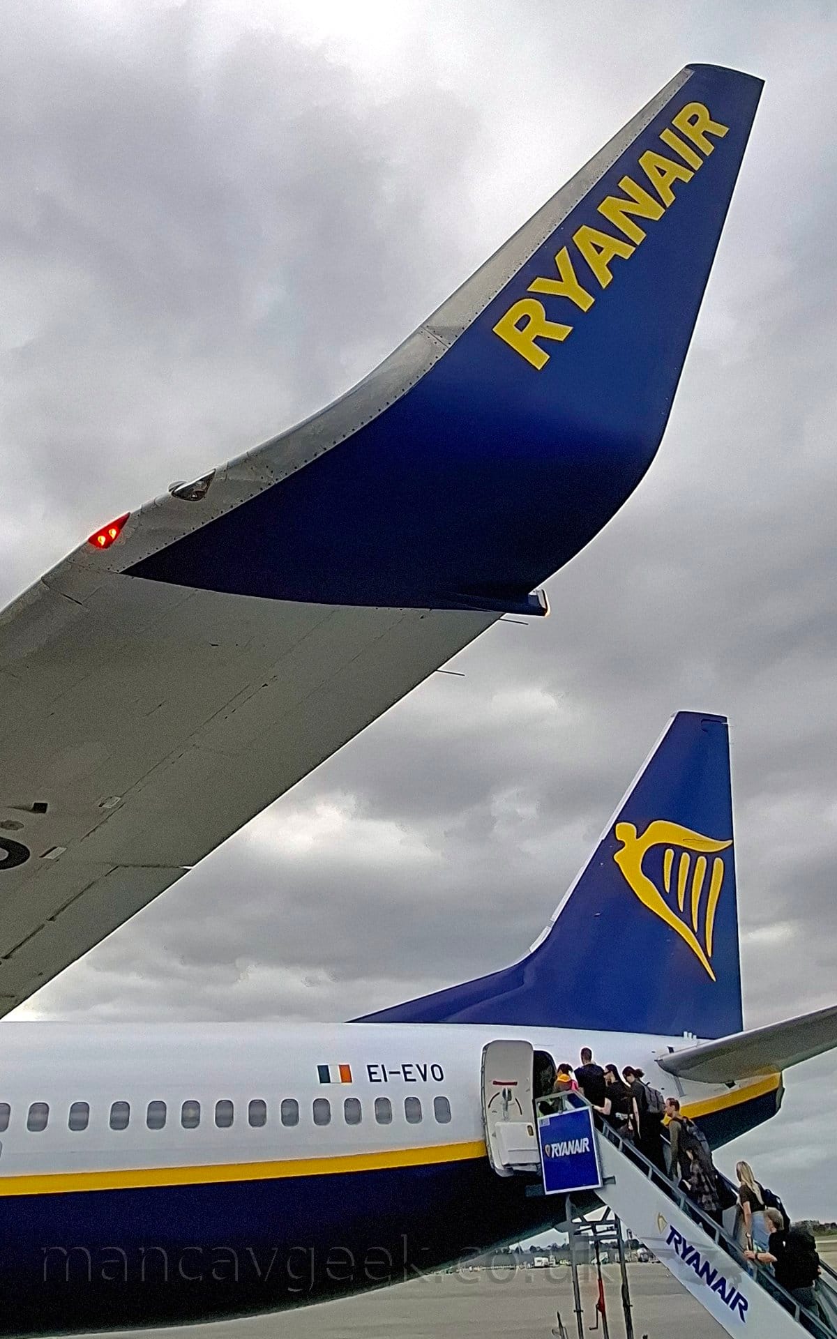 Close-up of the blue, up-turned wing-tip and rear fuselage of a jet airliner parked facing to the left. The plane has a white upper body, with a yellow stripe separating that from the dark blue belly, the black registration "EI-EVO" on the upper rear fuselage next to a small Irish flag. The tail is dark blue, with a large, yellow winged Irish harp in the middle. At the top of the frame, the planes wingtip is dark blue, with yellow "RyanAir" text. A white set of mobile airstairs with blue "RyanAir" titles is parked at the open rear door, with a queue of people climbing to gain entry. Dramatic cloudy grey skies fill the rest of the frame.