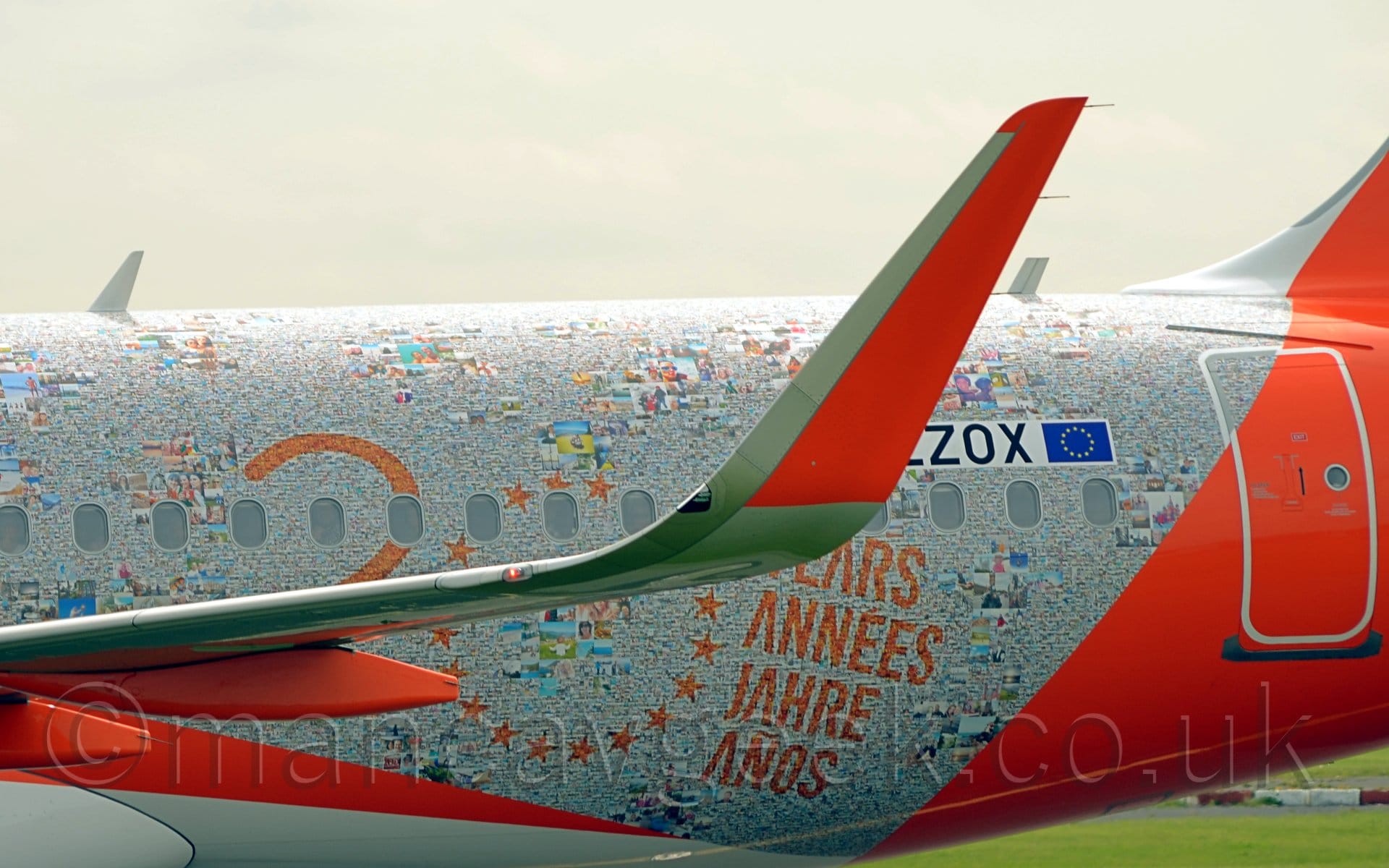 Close-up of the bright orange up-turned wing-tip of a jet airliner taxiing from right to left. In the background, the plane's body is covered in photos, some large, with easily distinguishable faces or locations, others are really small, with just hints of… something… there. Some of these are overlaid with orange text, first the number "20", with the "0" replaced with a ring of stars, obviously based on the stars in the EU flag, followed by various European words for "Years" stacked vertically - "Years", "Annees", "jahre", "anos". Thick orange stripe on the body mark the edges of the area covered by these photos. Green grass can be seen under the rear of the fuselage ion the background, and flat grey sky above completes the picture.