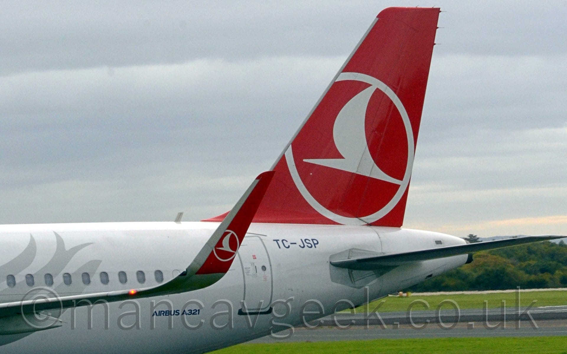 Close-up of the up-turned red wingtip of a white jet airliner taxiing from right to left. There is a silver flower on the planes body on the left of the frame. The wingtip has the white outline of circle, containing a white stylised flying bird, a logo repeated in a larger form on the tail. Green grass and trees can be seen in the background under the planes tail, with grey cloudy sky with additional streaks of grey cloud filling the rest of the frame.