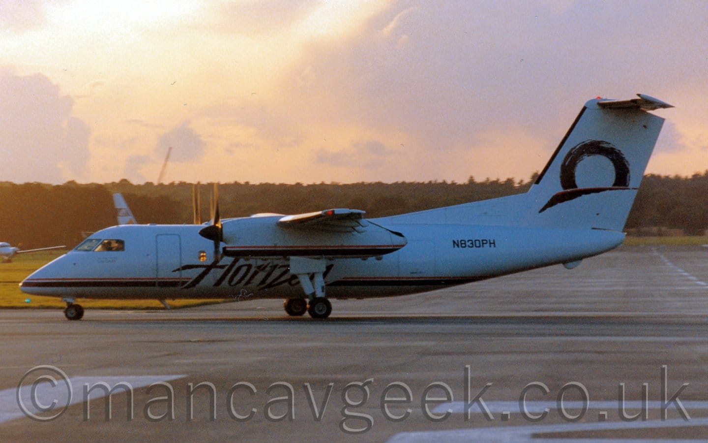 Heavily backlit side view of a high-winged, twin propellor-engined airliner parked facing to the left. The plane is almost entirely white, except for the black and red stripe running along the bottom of the fuselage, overlaid with black "Horizon" titles under the wings, and the partial black circle over a black brush stroke on the tail. A grey taxiway fills the foreground, extending off into the distance in the background on the right of the frame, with grass in the background on the left. Trees in the distance meet the orange sky, with purple-grey clouds, and a bright spot in the top left corner that is the sun which looks as if it will soon be setting.