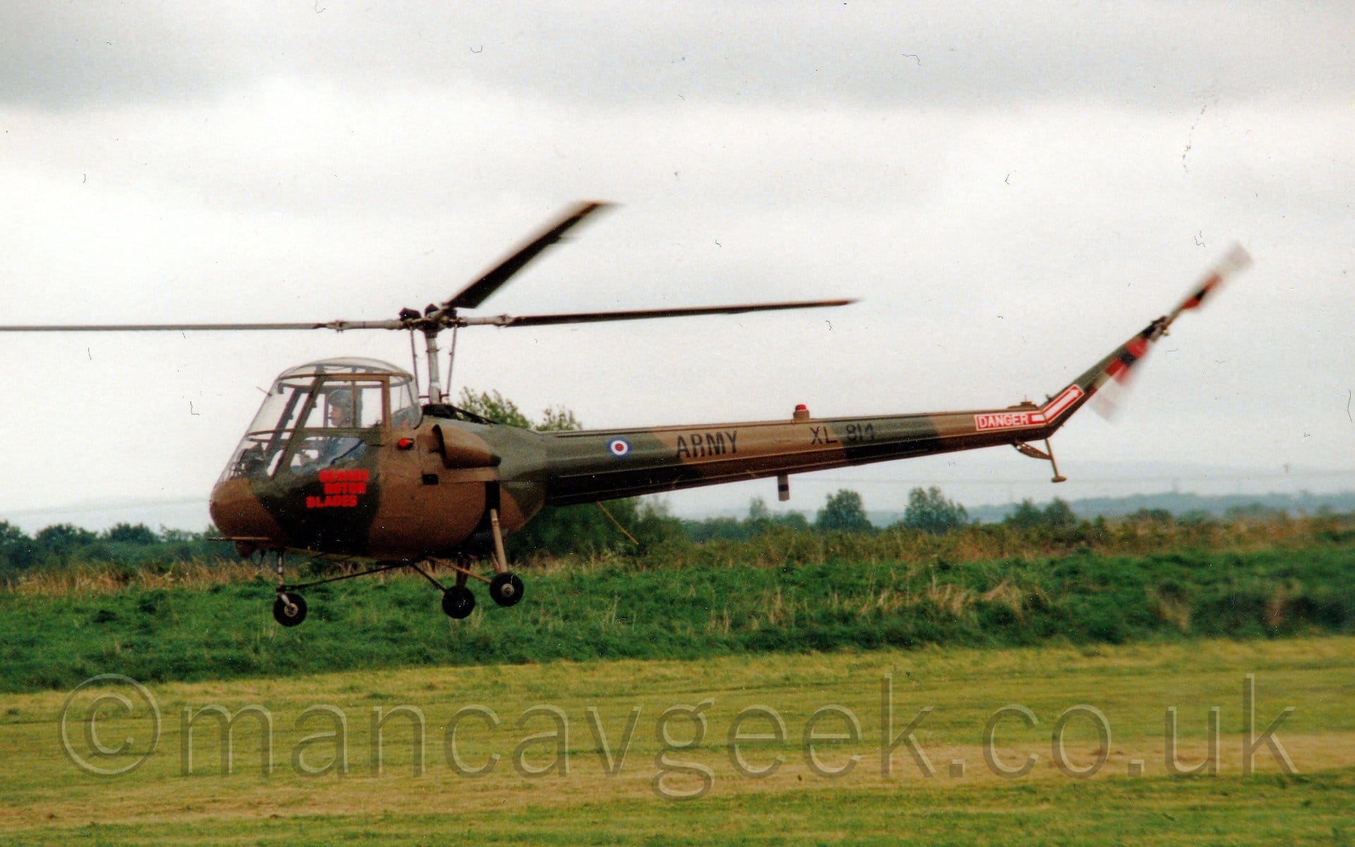 Side view of a small, rather ancient-looking helicopter flying from right to left above a grass airfield. The helicopter is covered in an olive green and brown camouflage scheme, with red "Beware rotor blades" text below the upturned-bucket cockpit. There is black "Army" titles and the serial "XL814" on the tail boom, as well as a red block with white text "Danger" and a white arrow pointing to the spinning tail rotor. It is flying just a few metres above a grass field, with bushes and trees in the distance, under flat grey sky.