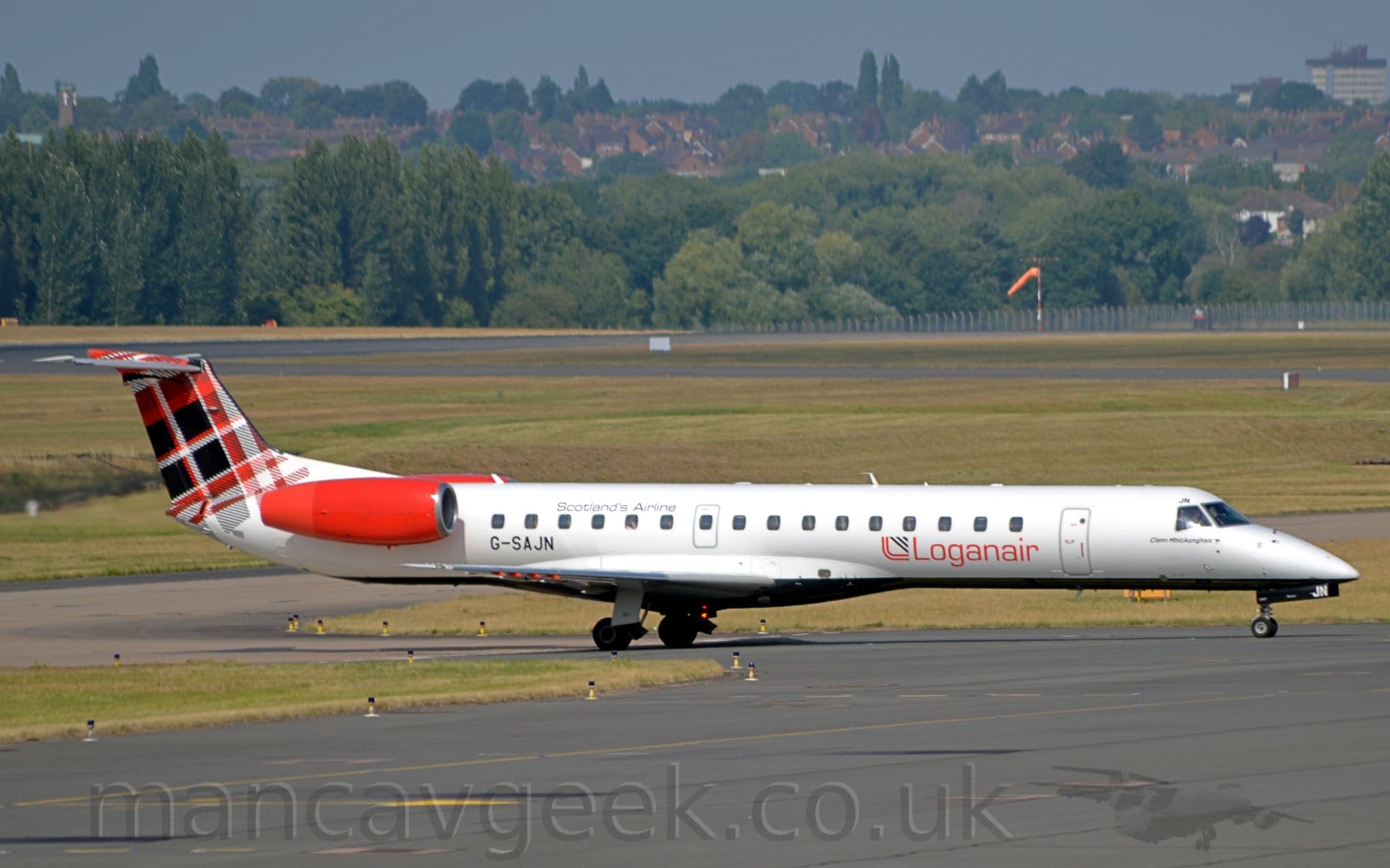 Side view of a twin engined jet airliner taxiing from left to right  along a black taxiway. The plane is mostly white, with a black belly. There are red "LoganAir" titles on the lower forward fuselage, with the black registration "G-SAJN" on the lower rear, just in front of and below the red engine pods mounted on the sides of the rear fuselage. The tail has a red, black, and white tartan pattern. Black taxiway fills the foreground, lined by green grass on the left. More grass, black taxiways, and a runway fill most of the background, leading up to trees in the distance, with houses nestled in the trees. Grey-blue sky fills the rest of the frame.
