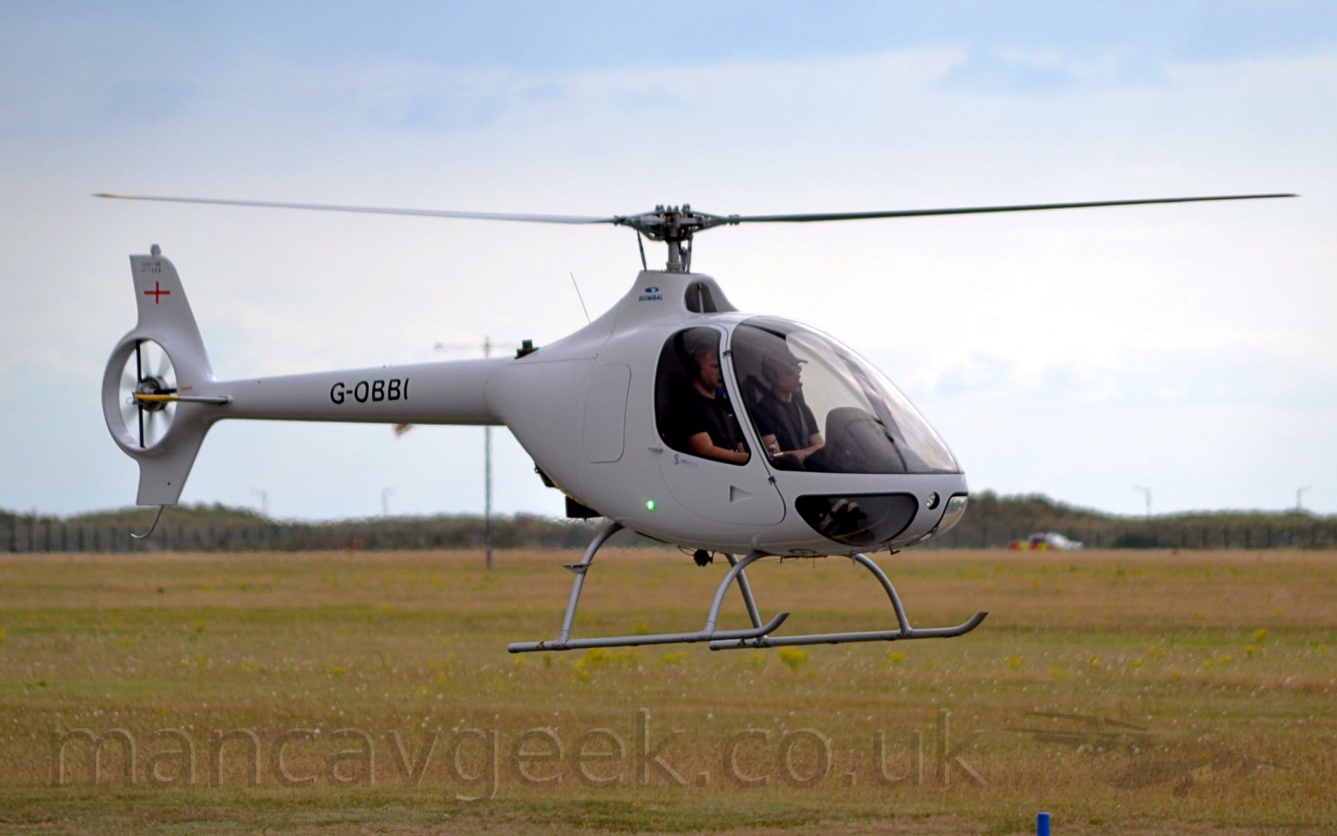Side view of a single engined light helicopter hovering a couple of metres above the ground. The helicopter is all white, with the black registration "G-OBBI" ion the tail boom. The tail-boom ends with a shrouded fan, buried in the tail. Two men wearing black t-shirts can be seen in the extensively glazed cockpit. Green grass in the background fills the bottom of the frame, while blue sky and sheets of white cloud fills the top.