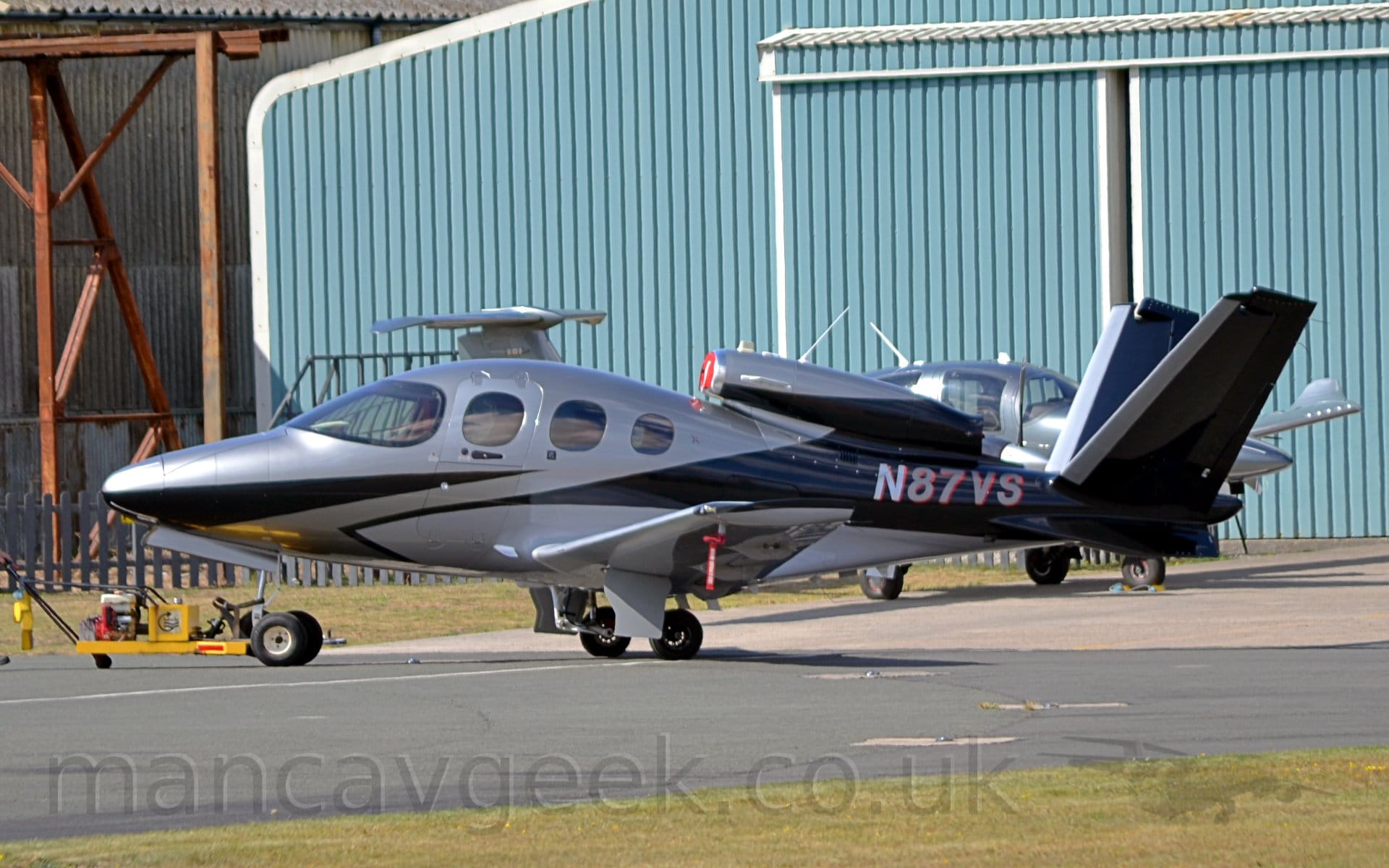 Side view of a single engined, very small BizJet parked on black tarmac facing to the left. The plane is mostly a silvery grey, with a slash of black on the nose, as well as a black rear fuselage and V-tail. A single engine is mounted above the sloping rear fuselage, exhausting between the V-tail. The silver registration "N87VS" is on the rear fuselage, under the rear of the engine. Black tarmac lined with green grass fills the foreground, with a large blue hangar with white trim fills most of the background. Another small, silvery plane is parked just in front of his hangar.