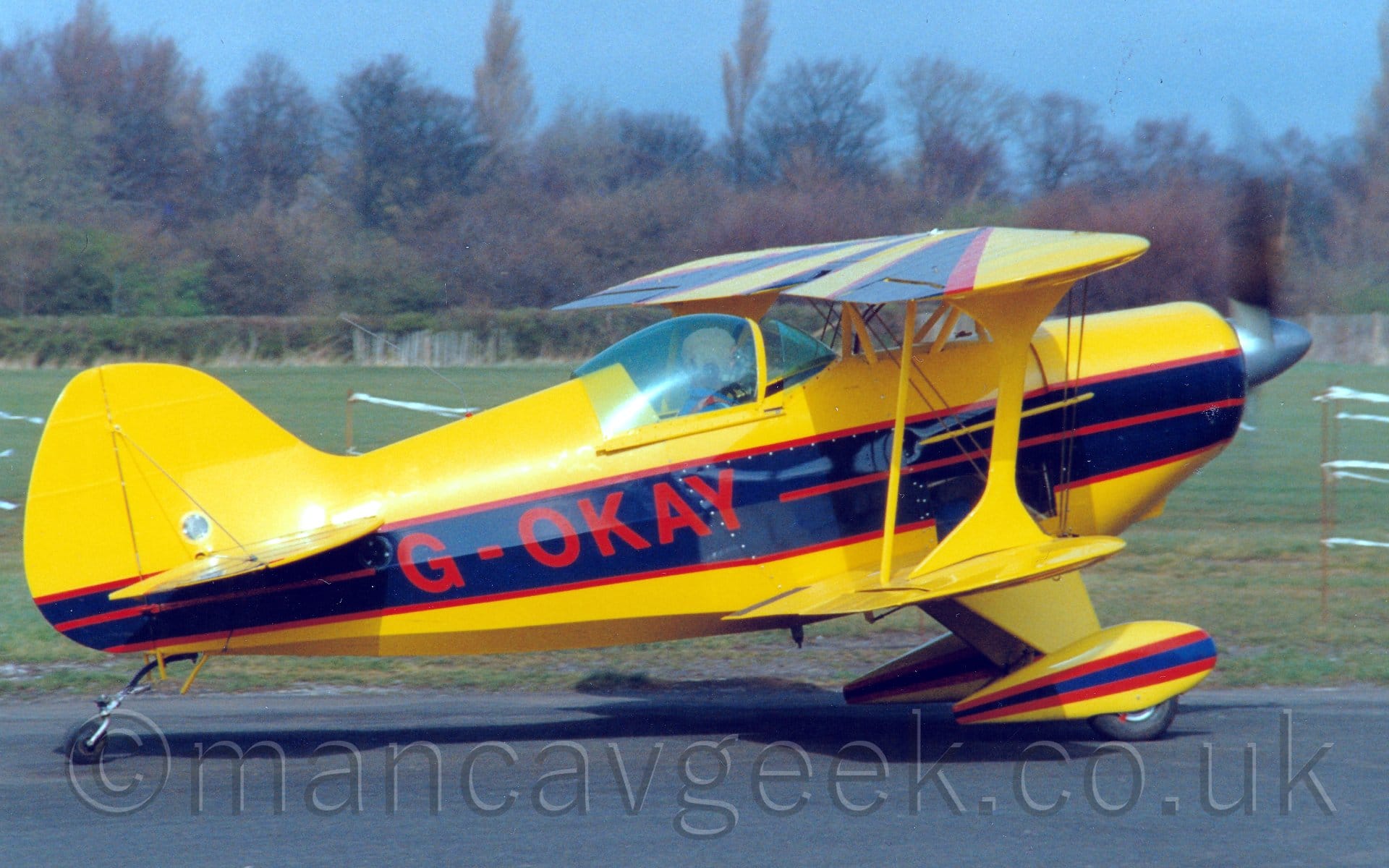 Side view of a single-engined, light biplane taxiing from left to right along a black taxiway. The plane is bright yellow, with a thick black stripe outlined in red running along the body, repeated on the spats covering the fixed undercarriage. T6he red registration "G-OKAY" is printed on the black stripe on the rear fuselage. More black stripes outlined in red are on the top of the top wing. The pilot is visible in the cockpit, wearing a cloth flying helmet and a blue shirt, seen through the blue-tinted canopy, which is slid slightly back. A large grassed area is in the background, leading up to trees and bushes in the distance, under blue skies.