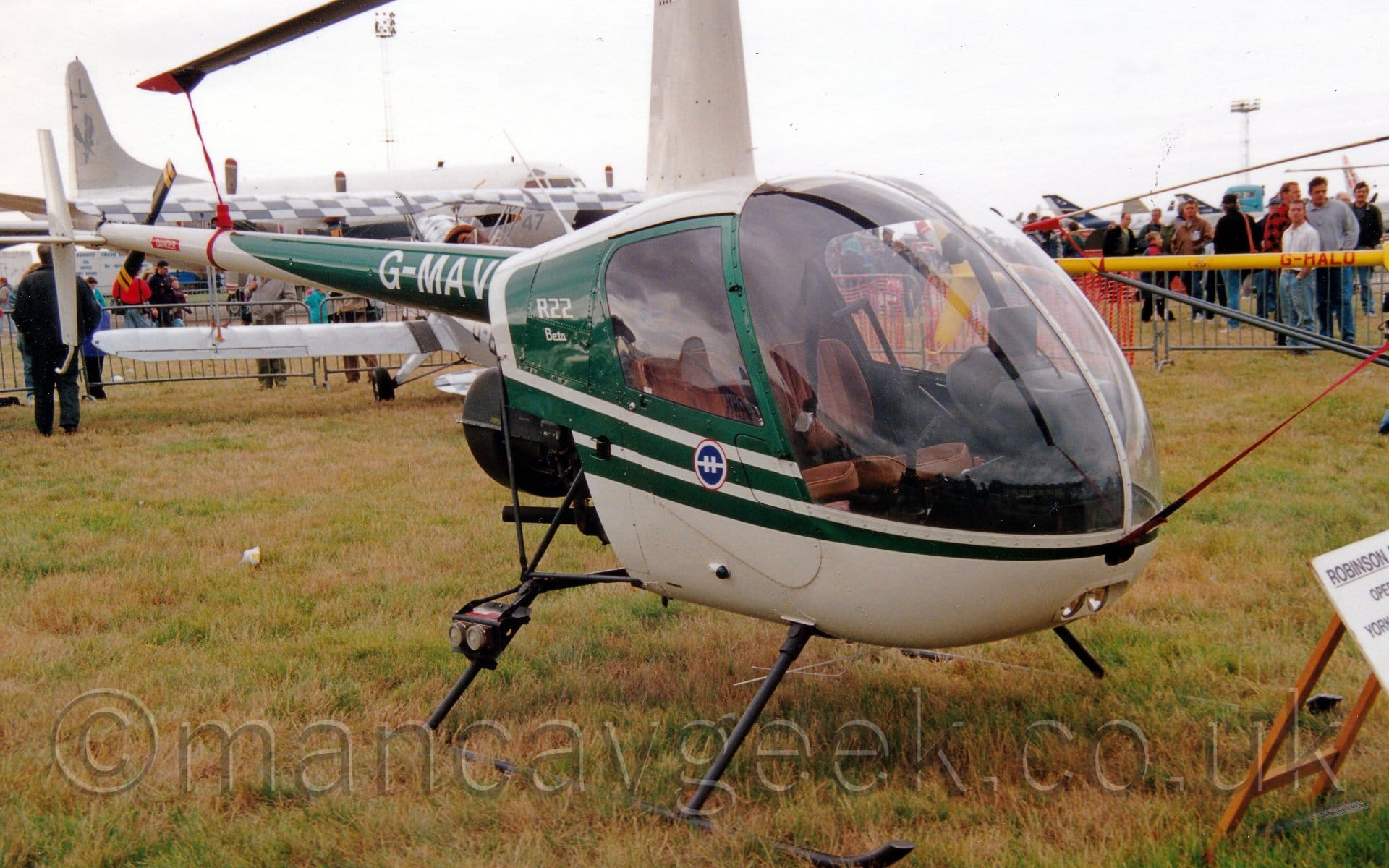 Side view of a single-engined light helicopter parked on grass facing to the right. The chopper is mostly white, with green stripes on the main body and tail boom. There is the white registration "G-MAVI" on the green part of the boom, and "R22 Beta" text on the green part of the body, just aft of the cockpit. A pair of brown seats can be see through the large, glazed canopy. In the background, a long, thin, yellow tail boom of a small helicopter can be seen, with the red registration "G-HALO" visible, is on the right of the frame. On the left of the background is a silvery-grey, single-engined bi-plane parked facing away from the camera, with a much larger, 4 propellor-engined military aircraft beyond that. Crowds are clustered around behind metal fences in the background, under grey skies.