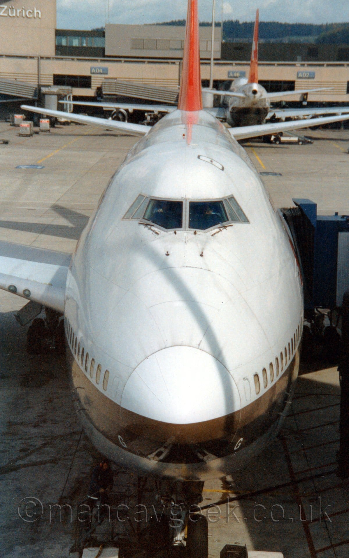 Vertically oriented head-on view of a very large, 4 engined jet airliner parked at the terminal, with the cockpit very slightly lower than the camera. The plane is mostly white, with a black and brown stripe wrapped around the nose and running towards the rear, while the tail is bright red. A blue airbridge is attached to the forward door on the lower left fuselage. An almost identical plane can be seen tail-on in the background, facing the sandy-brown terminal on the other side. On the left of the background, a sandy-brown tower with white "Zurich" titles is built on top of that terminal on the left. Rolling hills fill the background, slowly vanishing into haze in the distance.