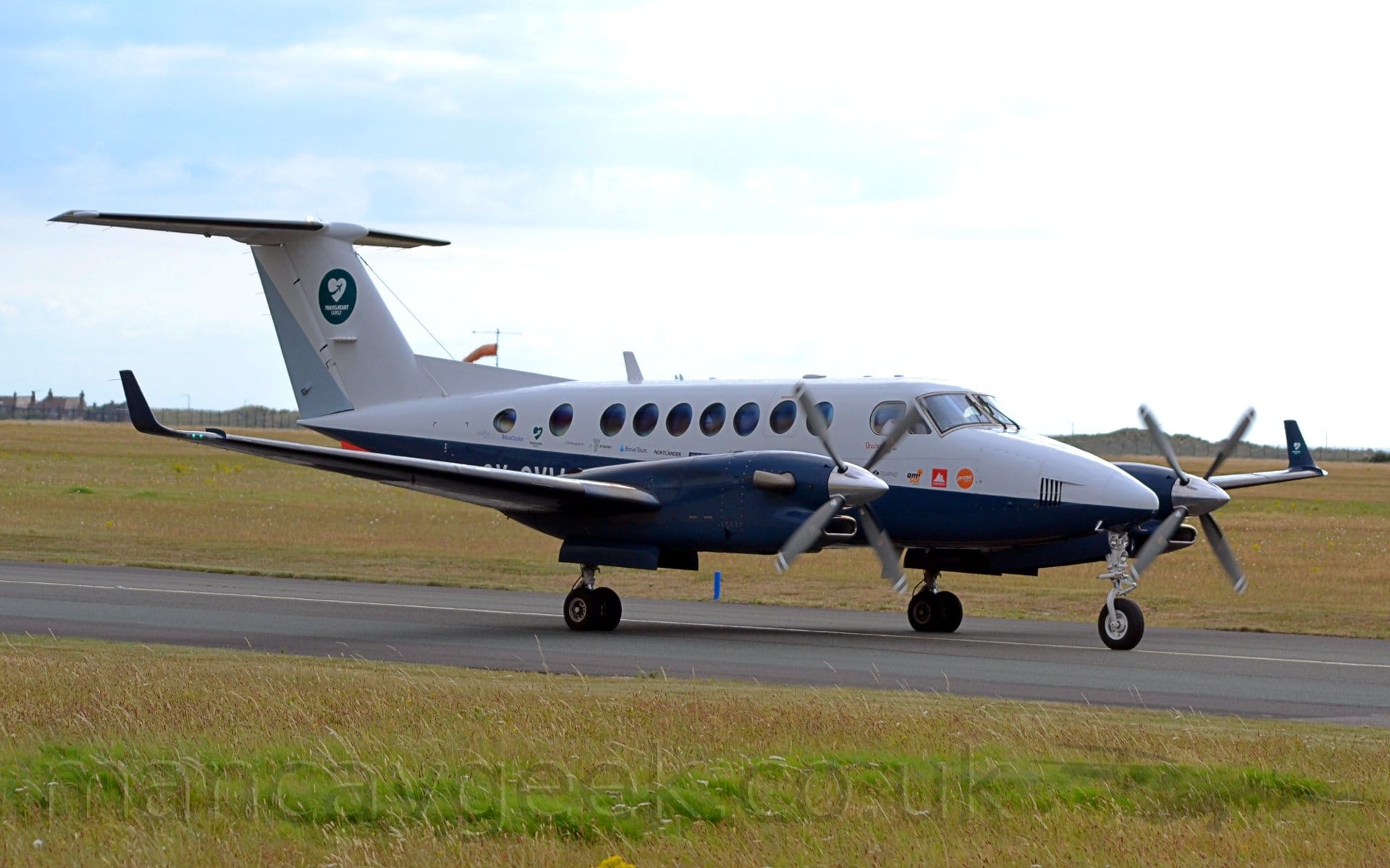 Side view of a twin-engined BizProp taxiing from left to right along a black tarmac taxiway. The plane is mostly white, with a dark blue belly and engine pods. The tail is white, with a green circle containing a white loveheart, itself containing a small image of a plane leaving a contrail. A similar logo (white loveheart with blue plane) is on the inside of the blue upturned wingtips. The engines have 4-bladed propellors spinning slowly at the front. Green grass lines the taxiway in the foreground, with more grass leading up to the horizon, under blue skies with bright white cloud.
