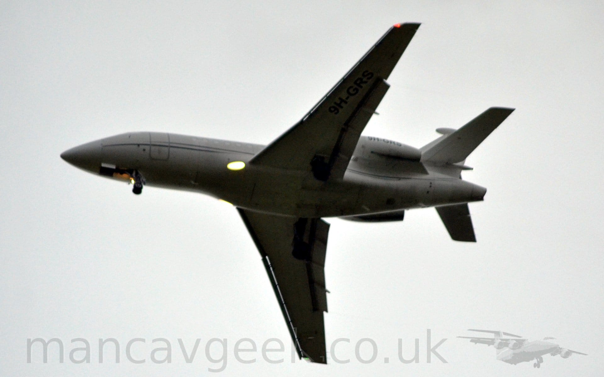 View from below of a 3 engined BizJet flying from right to left at a low altitude, with flaps extended and the undercarriage lowered, suggesting it is about to land. The plane is mostly white, with a black stripe swooping it's way along the lower fuselage, as well as along the tail. The black registration "9H-GRS" is on the sides of the engine pods mounted on the sides of the rear fuselage, as well as under the left wing. A bright light is shining in the wing root, slightly blowing out that part of the frame, as well as another light on the nose-wheel. Hazy grey sky fills the rest of the frame.