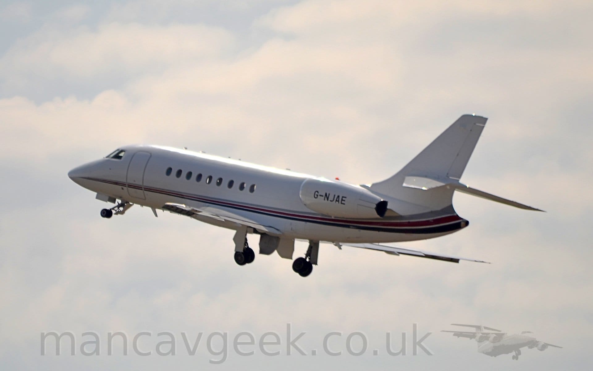 Side view of a twin engined BizJet flying from right to left at a low altitude, with the undercarriage in the process of being stowed away, flaps extended slightly from the rear of the wings, and the nose raised sharply, suggesting it has just taken off. The plane is mostly white, with  a grey, mauve, and blue stripe running along the body from the nose, getting wider as it moves rearwards and sweeping up into the lower reaches of the tail . The black registration "G-NJAE" is on the side of the engine pods, mounted on the sides of the rear fuselage. Strong sunlight is reflecting off the top of the fuselage Cloudy white sky with patches of blue fill the rest of the frame.
