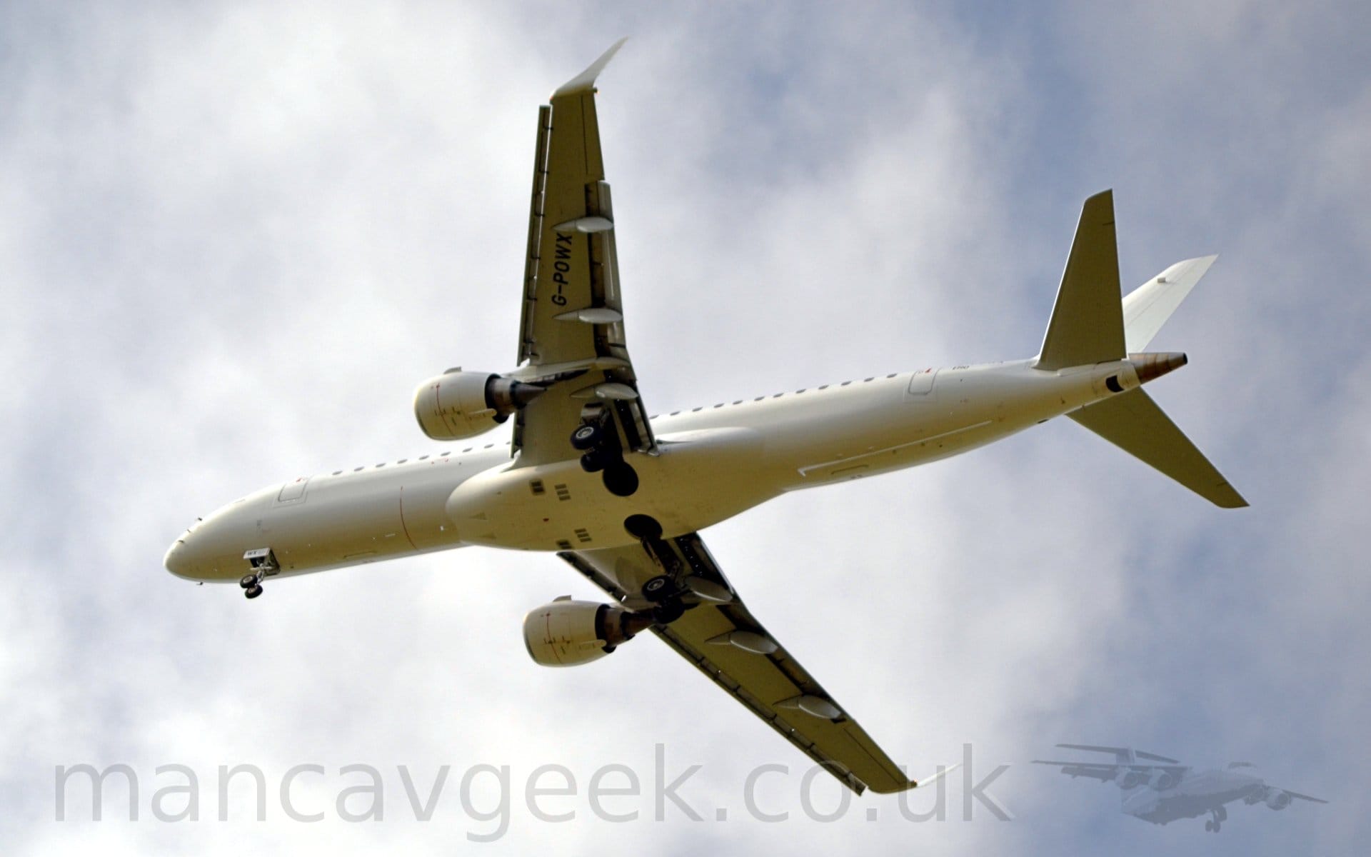 View from below of a twin engined jet airliner flying from right to left at a low altitude, with undercarriage lowered and flaps extended from the rear of the wings, suggesting that it is about to land. The plane is almost entirely white, with little more than the black registration "G-POWX" visible under the wings. Bright but hazy sky with blue patches threatening to break through fills the rest of the frame.