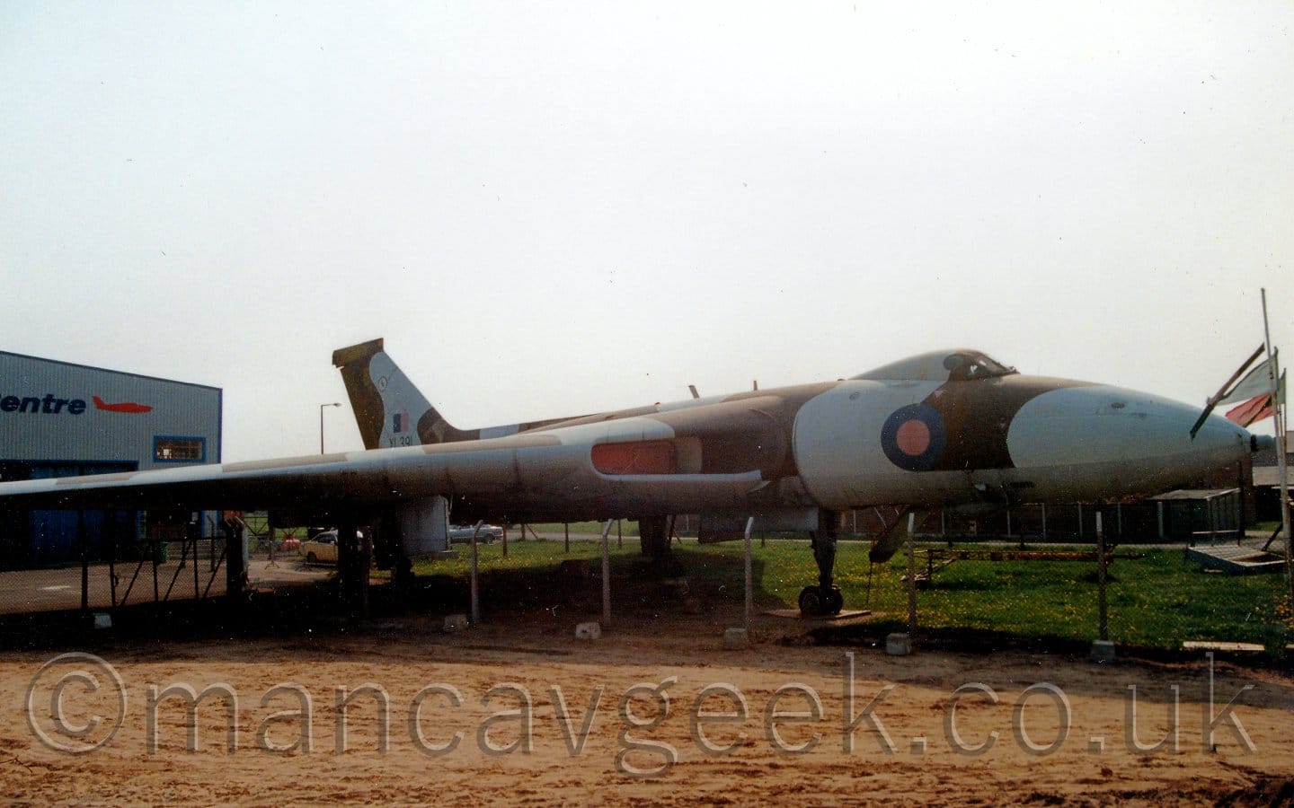 Somewhat backlit side view of a 4 engined, delta winged, military jet bomber aircraft parked facing to the right. The plane is covered in a grey and olive green camouflage scheme, with a blue and red roundel on the forward fuselage, under the large cockpit blister. The large air inlets in he wing root have big red baffles mounted inside, The black number 391 is on the tail, over a blue and red fin flash. A dirt field fills the foreground, with grass visible in the background under the plane. A grey building is on the left of the frame, behind the plane's wing. Bright but hazy sky fills the rest of the frame.
