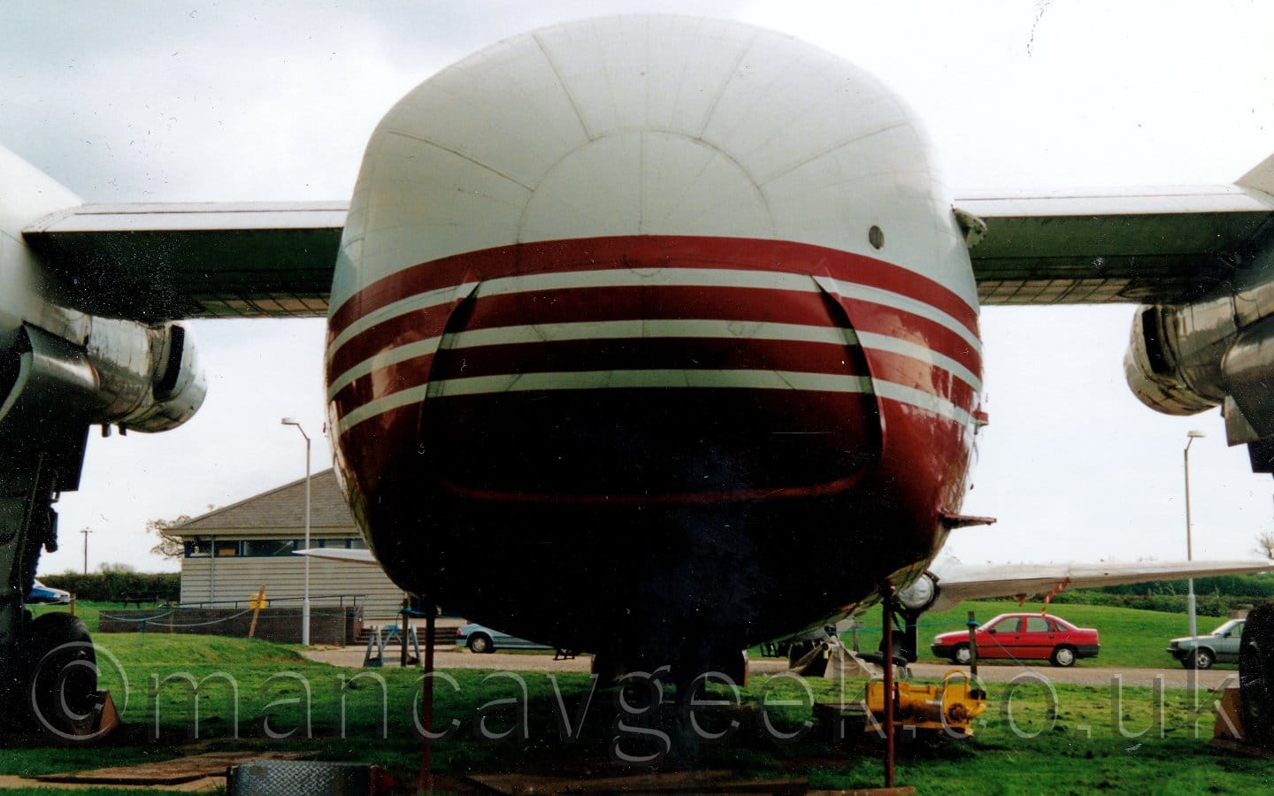 Rear view of a 4 propellor-engined cargo plane with a high-mounted wing and a very short body, parked on concrete pads surrounded by grass, facing away from the camera. The plane has white upper surfaces, with a red belly, and red stripes on the sides of the lower fuselage. The undercarriage can be seen suspended from the engine pods closest to the body. Green grass fills the foreground, with cars and another plane in the background, with more grass and a grey building in the distance, under grey skies.