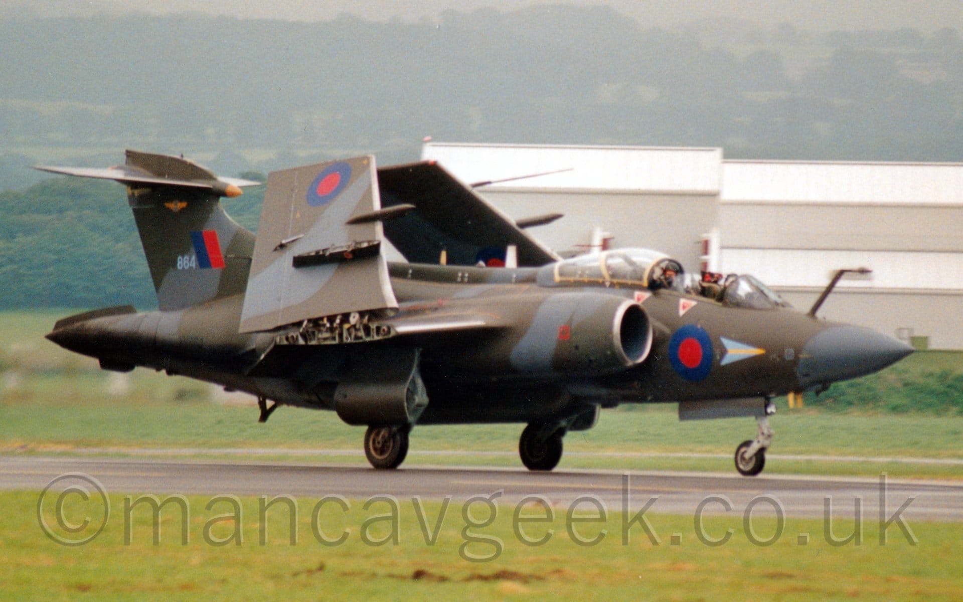 Side view of a twin jet-engined, military light bomber taxiing from left to right along a black runway with it's wings folded up. The plane is covered in an olive-green and grey camouflage, with blue and red roundels on the lower forward fuselage, under the cockpit, and under the outer wing panels, the latter visible because the wings are raised into their stowed position. There is a blue and red fin flash on the tail, next to the grey number "864". The cockpit canopy is in the slid back position, with both crew members visible in their tandem seats. Green grass lines the runway in the foreground, with a large grey building dominating the background on the right, with rolling, tree covered hills fading into the hazy grey sky.