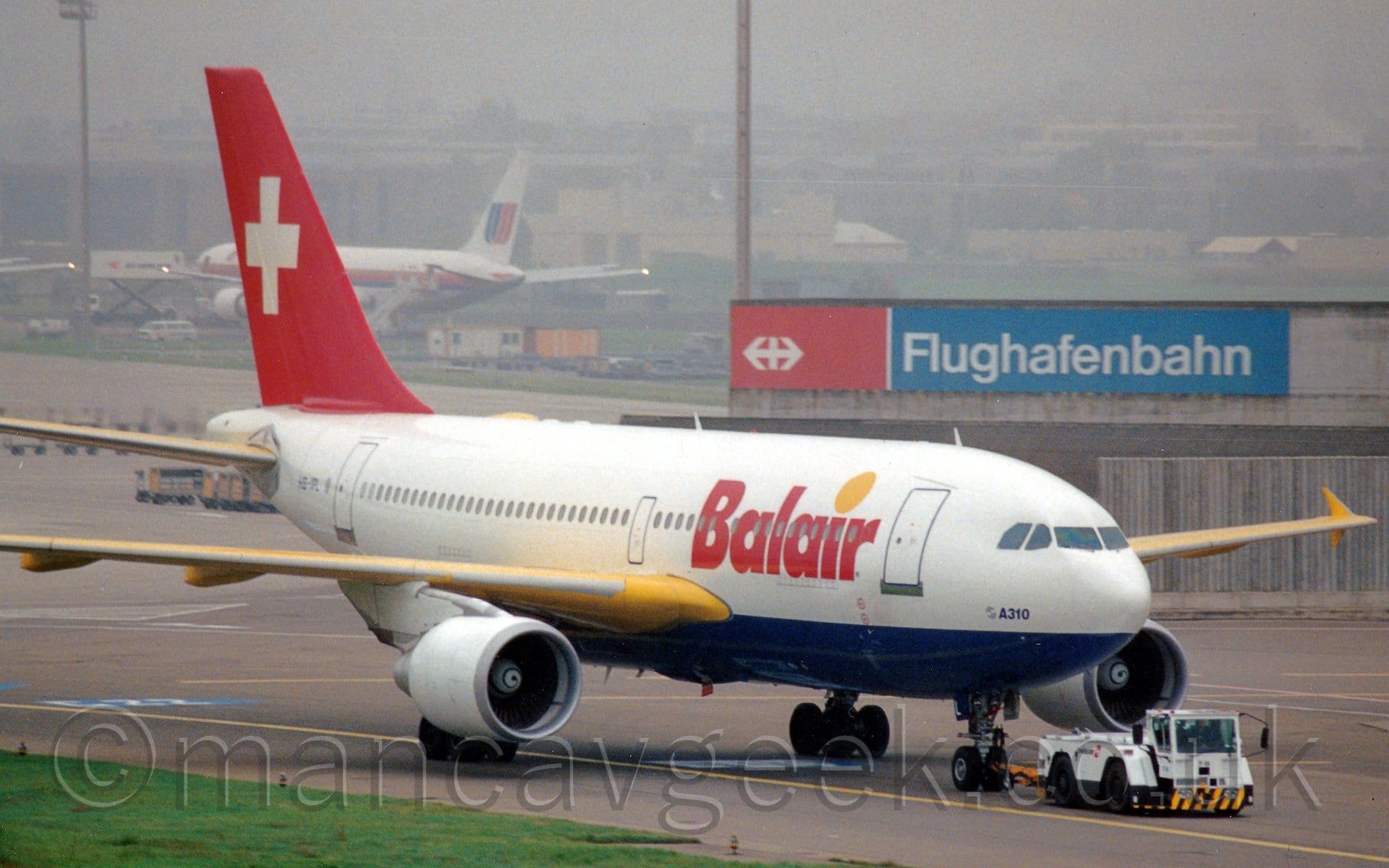 Side view of a twin engined jet airliner being towed from left to right by a white tug attached to the nosewheel by a long yellow pole. The plane is mostly white, with a dark blue belly, yellow wings, white engines, and a red tail with a large white cross in the middle. There are large red "BalAir" titles on the forward fuselage, with a large yellow dot above the lower-case letter "I". There are additional dark blue "A310" titles next to a circular blue Airbus logo on the nose under the cockpit windows. A grey concrete building with a red and blue banner with white "Flughafenbahn" titles is in the background on the right of the frame, with more buildings vanishing into haze in the distance, with a white jet airliner with an orange and blue stripe on the body parked in front.