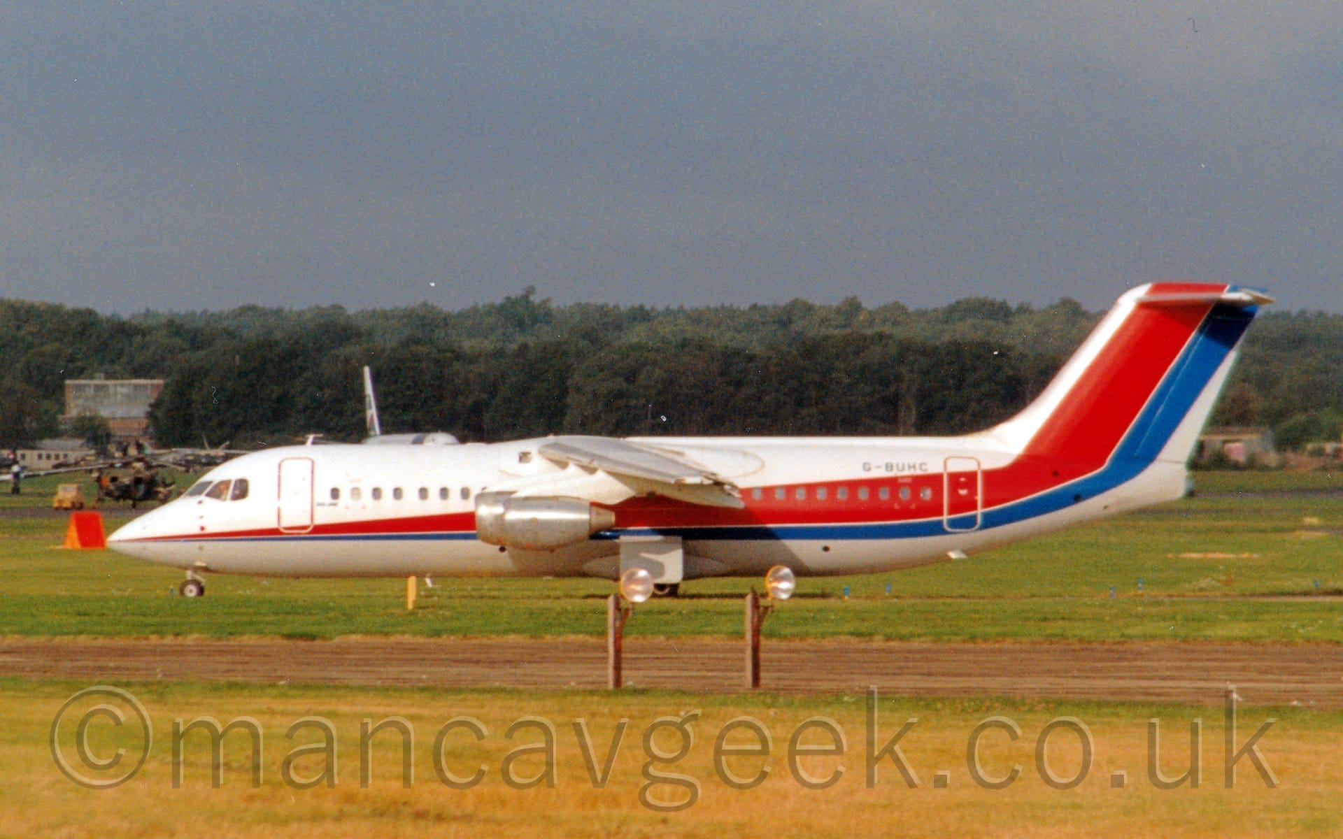 Side view of a high-winged, 4 engined jet airliner taxiing from right to left with flaps drooping behind the wings, suggesting it has just landed. The plane is mostly white, with a thin red and blue stripe running along the body, getting wider as it moves rearwards, before sweeping up to fill the tail. The blue registration "G-BUHC" . Yellowing grass fills the foreground at the bottom of the frame, with what appear to be lights on a pair of brown wooden poles next to a taxiway. Several other planes can be seen in the background behind this planes nose, with rows of trees in the distance under grey-blue sky.