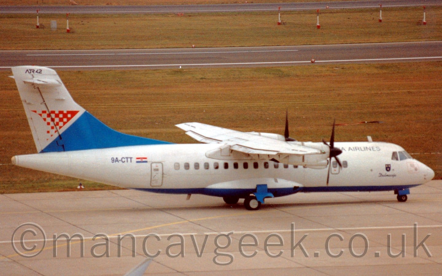Side view of a high-winged, twin propellor-engined airliner taxiing from left to right. The plane is mostly white, with a blue belly, and black "Croatia Airlines" titles on the upper forward fuselage. The black registration "9A-CTT" is on the upper rear fuselage, under the blue and white tail which has a triangular red logo. Grey taxiway fills the foreground, with yellowing grass either side of taxiways in the background.