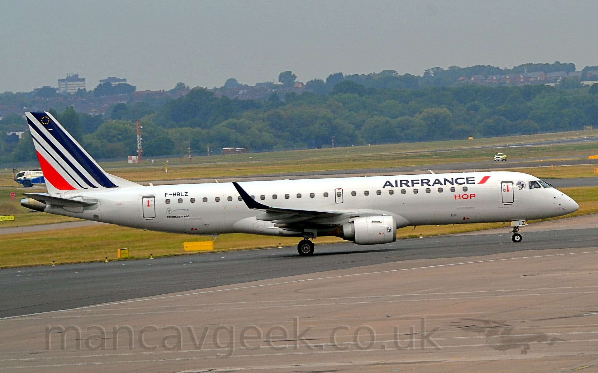 Side view of a twin engined jet airliner taxiing from left to right. The plane is mostly white, with blue "Air France" titles on the upper forward fuselage, and smaller "Hop" titles underneath. The black registration "F-HBLZ" is on the upper rear fuselage, while the last 2 letters "LZ" are on the open nose-wheel doors. 3 diagonal blue stripes are on the tail, getting narrower the further back they are, with a fourth, red stripe at the back, the same width as that at the front. Grey concrete apron and black concrete taxiway fill the foreground, with grass, taxiways, and a runway in the background, leading up to trees in the distance, under grey skies.