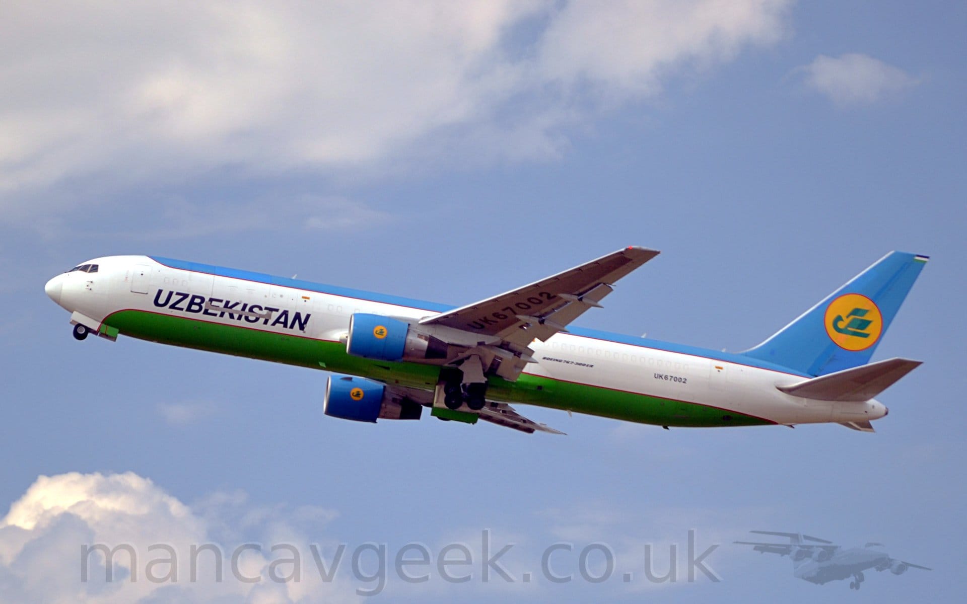 Side view of a twin engined jet freighter howling into the air, flying from right to left with the undercarriage being stowed, flaps extended from the rear of the wings, and the nose raised significantly, suggesting it has just taken off. The plane has a wide white stripe running along the middle of the body, with blue on the upper body and tail, and green on the belly, both outlined in red. There are black "Uzbekistan" titles on the lower forward fuselage, and the registration "UK67002" on the lower rear, with additional black text "Boeing 767-300ER" just aft of the wing root. On the tail, a stylised green flying bird is in the middle of a yellow circle that is outlined in red. Pale blue sky fills most of the rest of the frame, with the plane threading it's way between fluffs of white cloud at top and bottom.