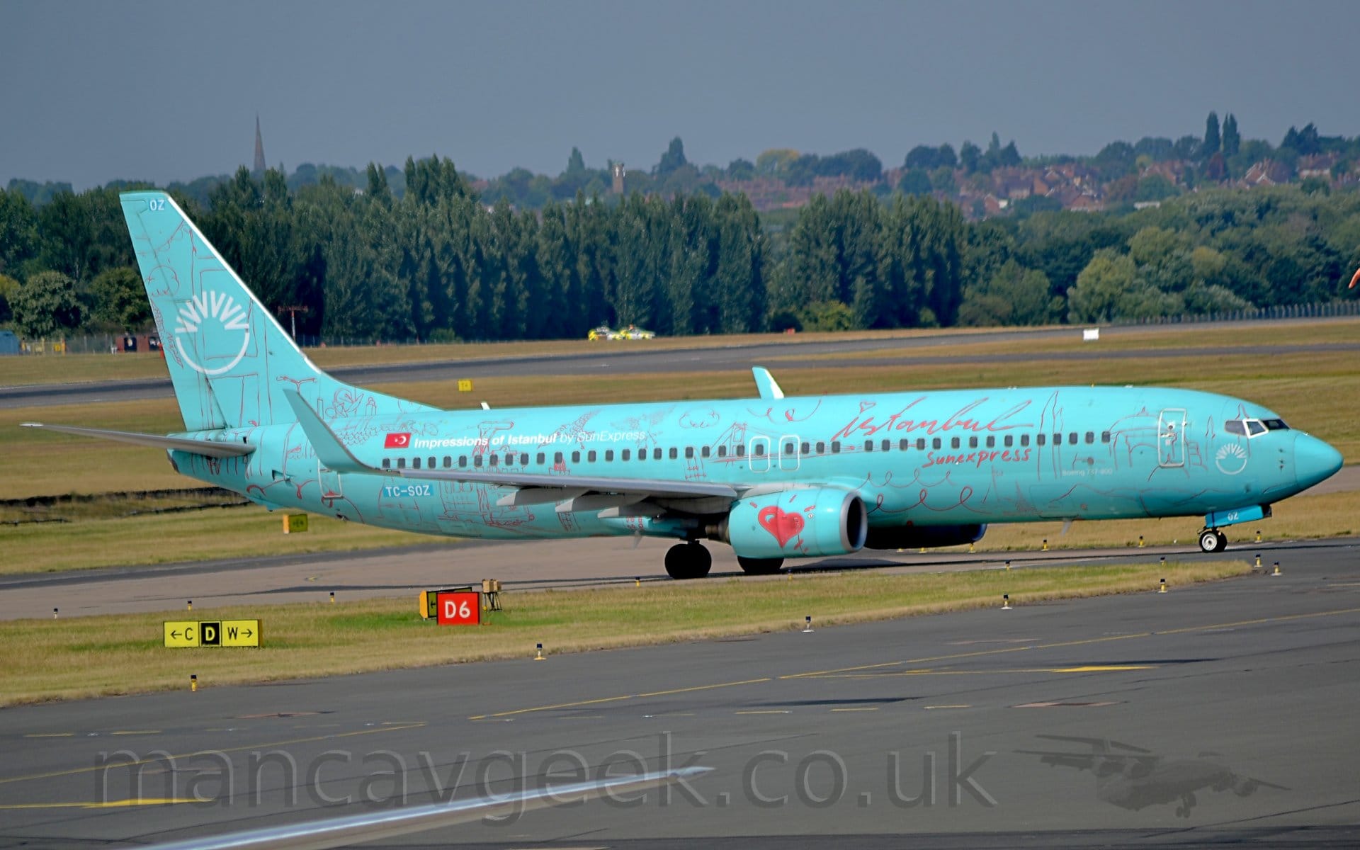 Side view of a twin engined jet airliner taxiing from left to right. The plane is a very pale blue, with red, seemingly hand-written "Istanbul" titles on the upper forward fuselage, above the forward cabin windows, with additional "SunExpress" below the windows. Hand-drawn red images of buildings, bridges, vegetation, weather, and curly squiggles cover the rest of the airframe, with a large red love-heart on the side of the engine pods. There is white text "Impressions of Instanbul by SunExpress" next to a small Turkish flag on the upper rear fuselage. A white semi-circle topped with 6 white chevrons themselves arranged in a semi-circle, while a smaller version of this logo appears on the nose. A black taxiway fills the foreground, with grass, more taxiways, and a runway in the background lead up to trees in the distance, with houses beyond that, under grey sky.