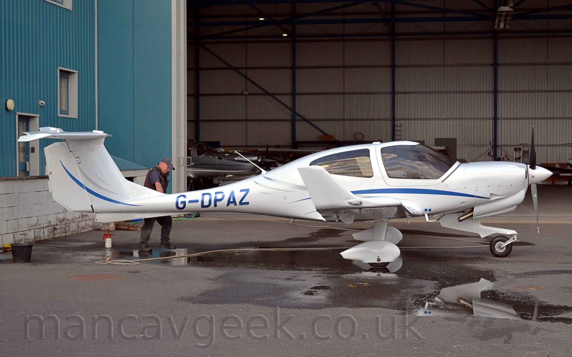 Side view of a single-engined light aircraft parked on black tarmac facing to the right. The plane is mostly white, with a blue stripe swooping it's way along the body, the blue registration "G-DPAZ" on the rather thin rear fuselage. White spats cover the fixed main landing gear, while the tips of the wings are turned upwards. A large wet patch covers part of the tarmac under the plane, where it has been washed. A man wearing a grey cap and a grey jacket over a blue t-shirt can be seen on the far side of the planes tail. In the background, a blue hangar with white trim stretches across the frame, with the open door revealing the white inner walls, and a couple of planes parked inside.