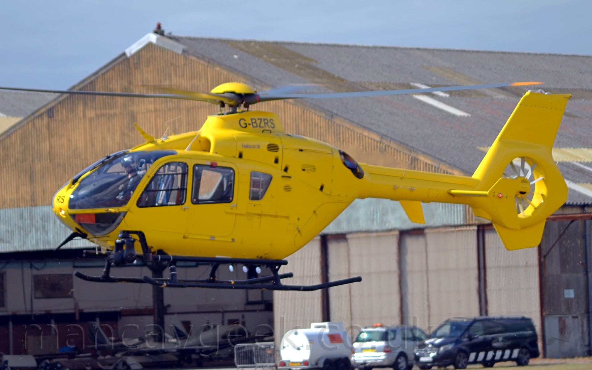 Side view of a helicopter hovering several metres above the ground at an airport. The copter is almost entirely bright yellow, with only the black registration "G-BZRS" on the engine cowling at the top of the fuselage, under the rotors, with smaller text "Babcock" under that, and the letters "RS" on the nose. A large grey hangar with a faded yellow roof stretches across the background, with several vehicles parked in front on the right of the frame, and several planes visible through the wide open doors on the left. Pale blue sky can be seen over the top of the hangar at the top of the frame.
