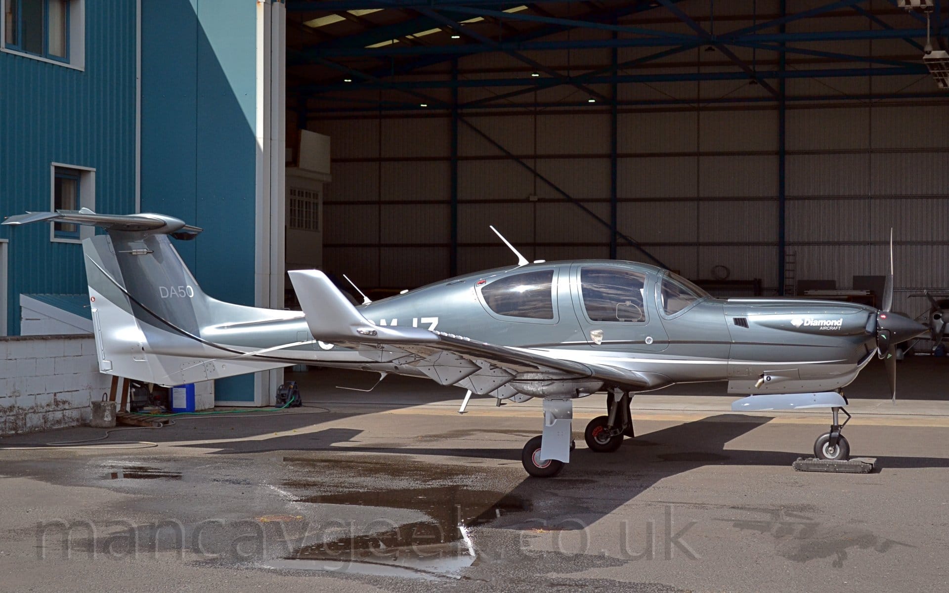 Side view of a very sleek, single-engined light aircraft parked on concrete apron facing to the right, that looks as if it is moving fast even when it is standing still. The plane is mostly a silvery grey with a white belly, and the white registration "G-CMJZ" on the side of the rear fuselage, partially obstructed by the right wing. Additional white text "DA50" is on the tail, and "Diamond Aircraft" on the engine cowling on the nose. In the foreground, a large wet patch with puddle is on the ground on the left, although the rest of the ground is bone dry. The plane is casting a strong shadow on the ground, with sunlight coming from just left of overhead. The planes nose-wheel is resting on what looks like a black plastic box, but is actually a very small, remotely controlled tug. In the background, a large blue hangar with white trim fills the frame, it's doors it's doors wide open, with a couple of residents visible inside.