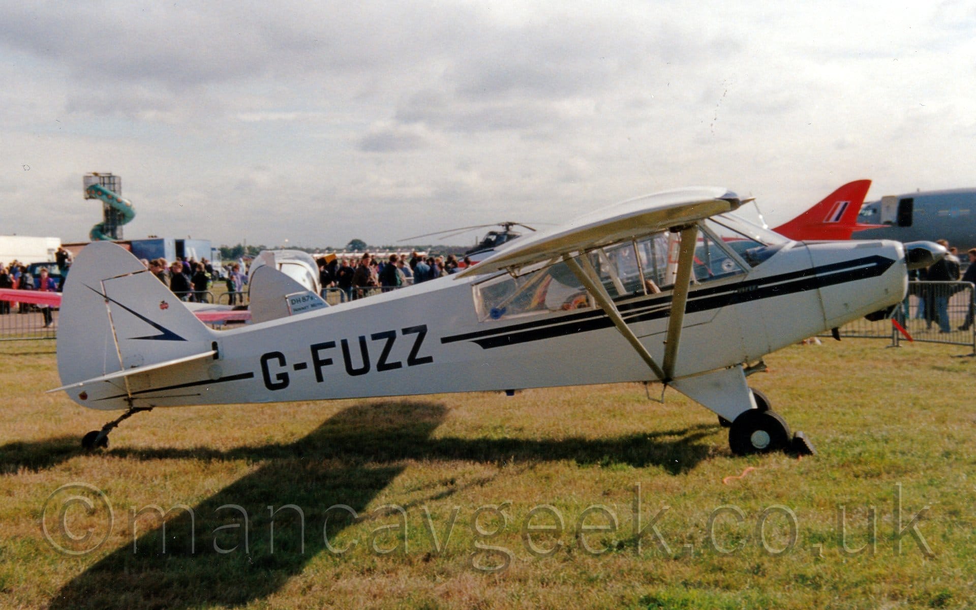 Side view of a white, high-winged, single-engined light aircraft parked on grass facing to the right. The plane has a blue stripe running along the body from just behind the propellor in the nose, overlaid with the blue registration "G-FUZZ". The plane is casting a strong shadow on the grass, the shadow of the wing suggests that the sun is fairly low down on the right. Green grass fills the foreground, with some other planes visible in the background, some surrounded by crowds of people. Bright but hazy sky with fluffs of white sky fill the rest of the frame.