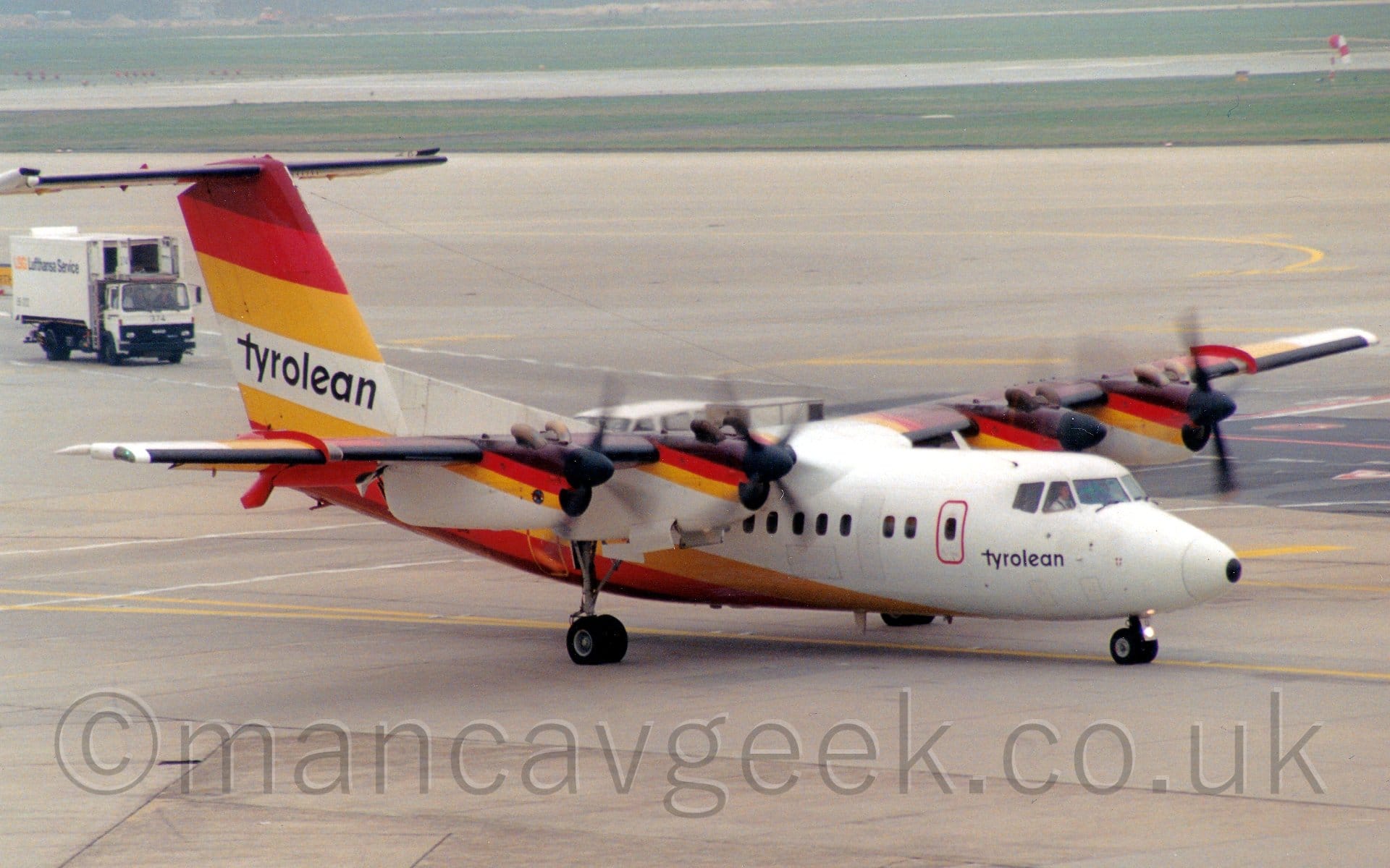 Side view of a high-winged, 4 propellor-engined airliner taxiing from left to right. The plane is mostly white, with thick yellow, red and brown stripes on the lower rear fuselage, angled up to the lower part of the tail, with a reflected version of this stripe on the upper part of the tail, as well as on the upper forward sections of the engine pods. There are small, black, "Tyrolean" titles on the forward fuselage, under the cockpit windows, with a larger version on the white portion of the tail. Grey concrete apron fills the foreground and most of the background, with bands of green grass bordering runways in the distance. A white airport truck can be seen in the background on the left, following a roadway marked out with white lines on a taxiway.