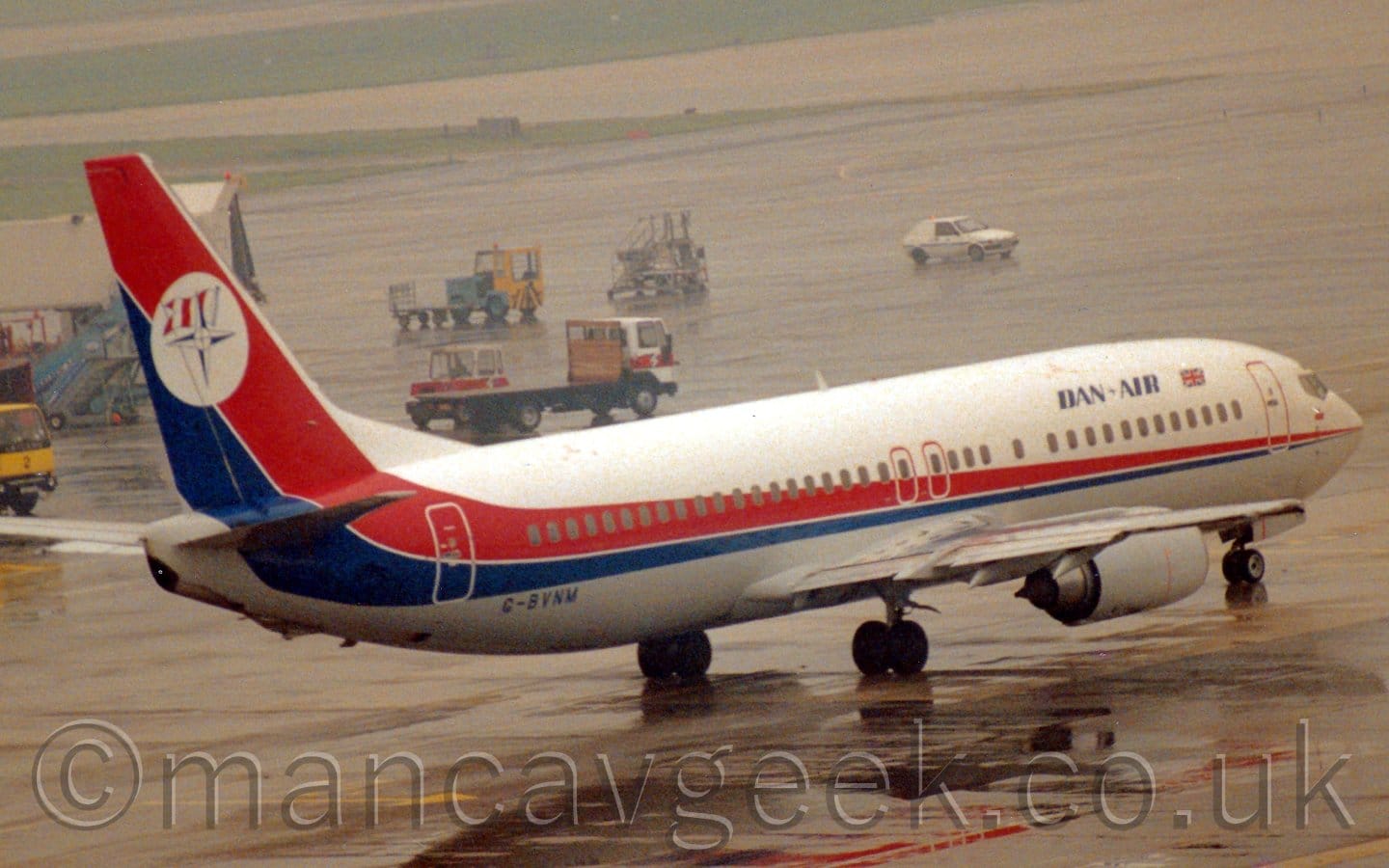 Somewhat grainy side view of a twin engined jet airliner taxiing from left to right on a rainy day. The plane is mostly white, with a red and blue stripe running along the body, getting wider as it moves rearwards, until it sweeps up to fill the tail. There are blue "Dan Air London" titles on the upper forward fuselage, next to a small British flag, and the blue registration "G-BVNM" on the lower rear. The tail has a white circle, with a blue compass star in the middle, and flag with 3 red and white vertical stripes behind. Wet apron fills most of the frame, with several airport vehicles moving around in the background, and a couple of wide strips of grass lining taxiways in the distance.