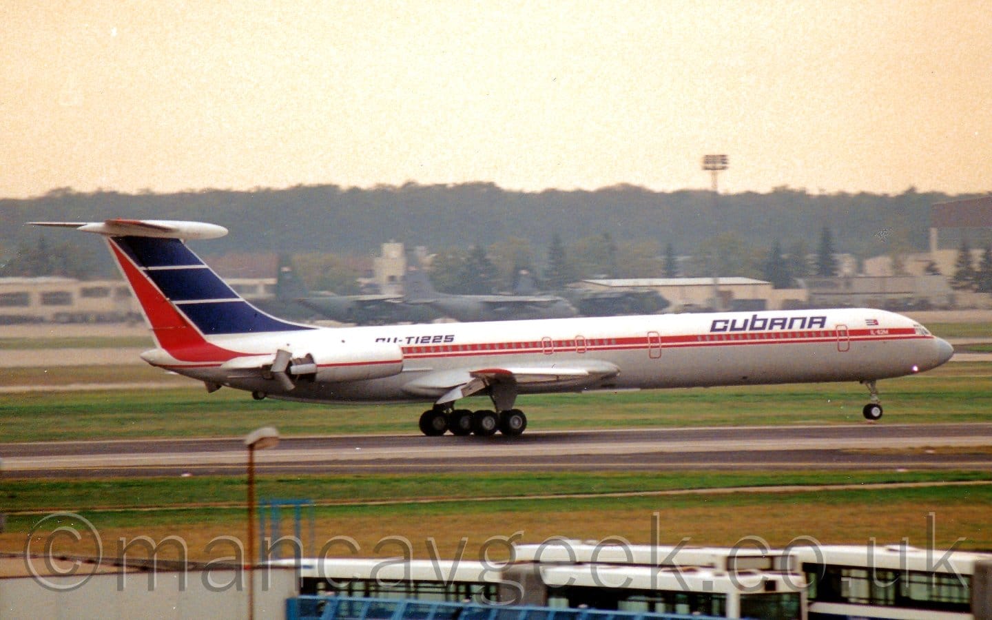 Side view of a 4-engined jet airliner moving from left to right at high speed. The plane has it's nose-wheel in the air while the main wheels are on the ground, flaps extended from the rear of the wings, and thrust reverser buckets deployed behind the engines mounted in pairs on the sides of the rear fuselage, suggesting the plane has just landed. There is a red stripe running along the body, turning up to fill the rear of the tail., and blue "Cubana" titles on the upper forward fuselage, with the black registration "CU-T1225" on the upper rear fuselage, partially hidden behind the engine pods. Green grass fills the foreground, with several buses parked right at the bottom of the frame. More grass on the other side of the runway leads up to a group of grey and green military transport planes parked in front of cream-coloured hangars. Trees in the distance slowly vanish into the bright but hazy golden sky.