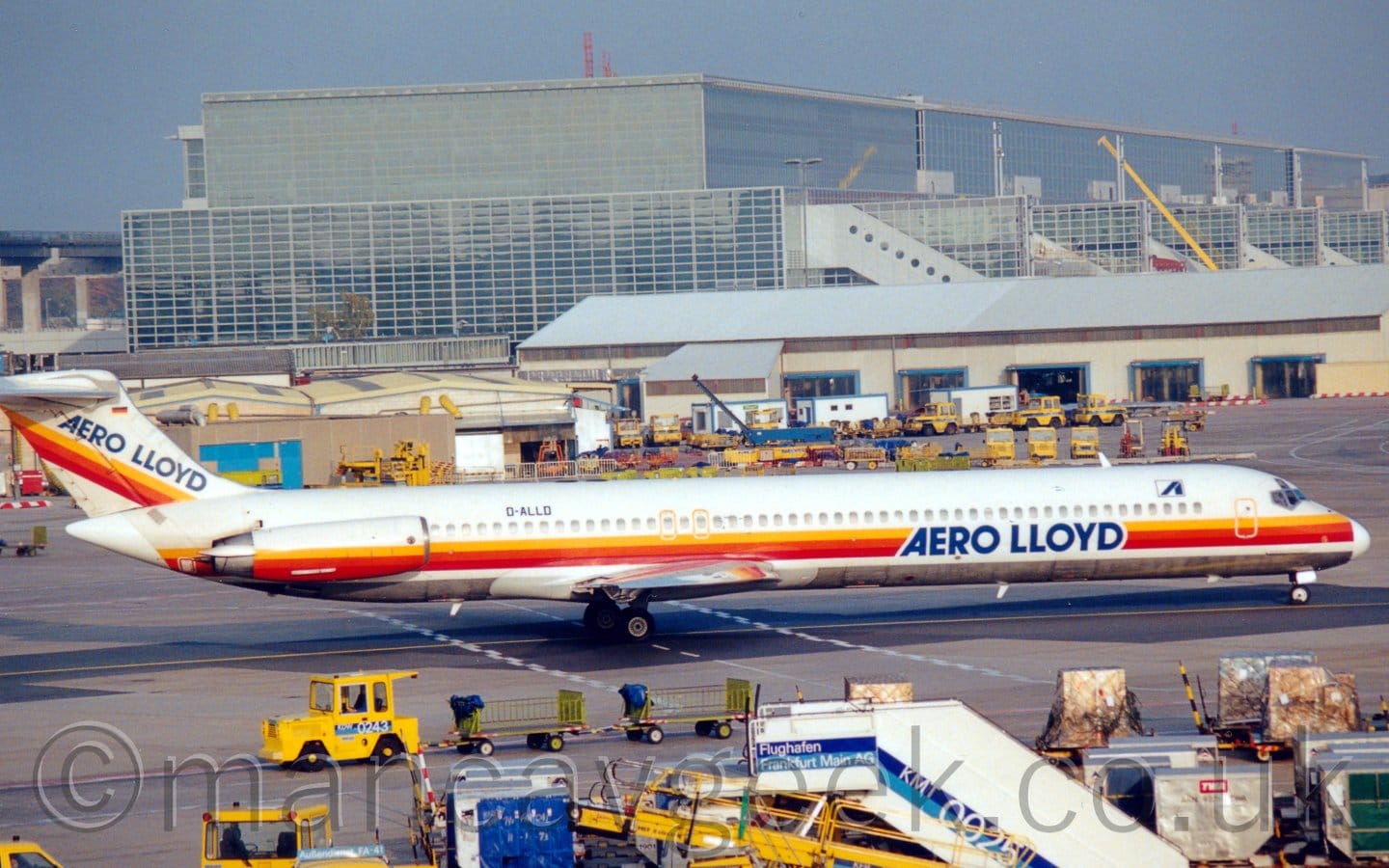 Side view of a twin engined jet airliner with the engines mounted on the sides of the rear fuselage, taxiing from left to right. The plane is mostly white, with a yellow, orange, and red stripe running along the body under the passenger cabin windows, overlaid with blue "Aero Lloyd" titles on the lower forward fuselage. A similar stripe runs across the engine pods, and diagonally up the tail, where there are more "Aero LLoyd" titles. Metal luggage containers, white mobile airstairs, and yellow baggage loading belts litter the foreground, along with some other airport furniture, while a large, black and grey terminal stretches across the frame in the background, looming over everything else. Blue sky fills the rest of the frame.