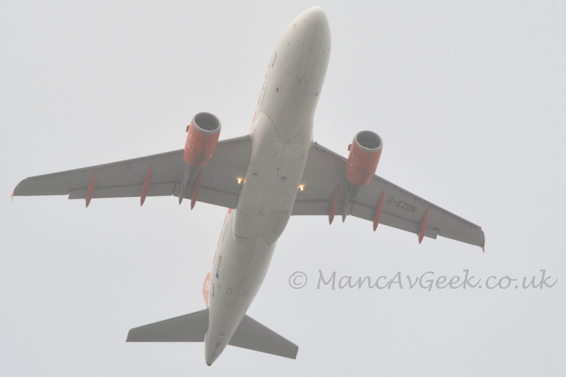 View from below of a twin engined jet airliner flying from the bottom to the top of the frame, with undercarriage already tucked away, flaps extended from the rear of the wings, and the nose raised sharply, suggesting it has just taken off. The plane is almost entirely white, with indistinct orange text on the side of the fuselage, orange engine pods mounted under the wings, and orange covers over the flap tracks under the rear of the wings. A pair of bright lights are mounted in the wing roots, facing forwards. Flat grey sky fills the rest of the frame.