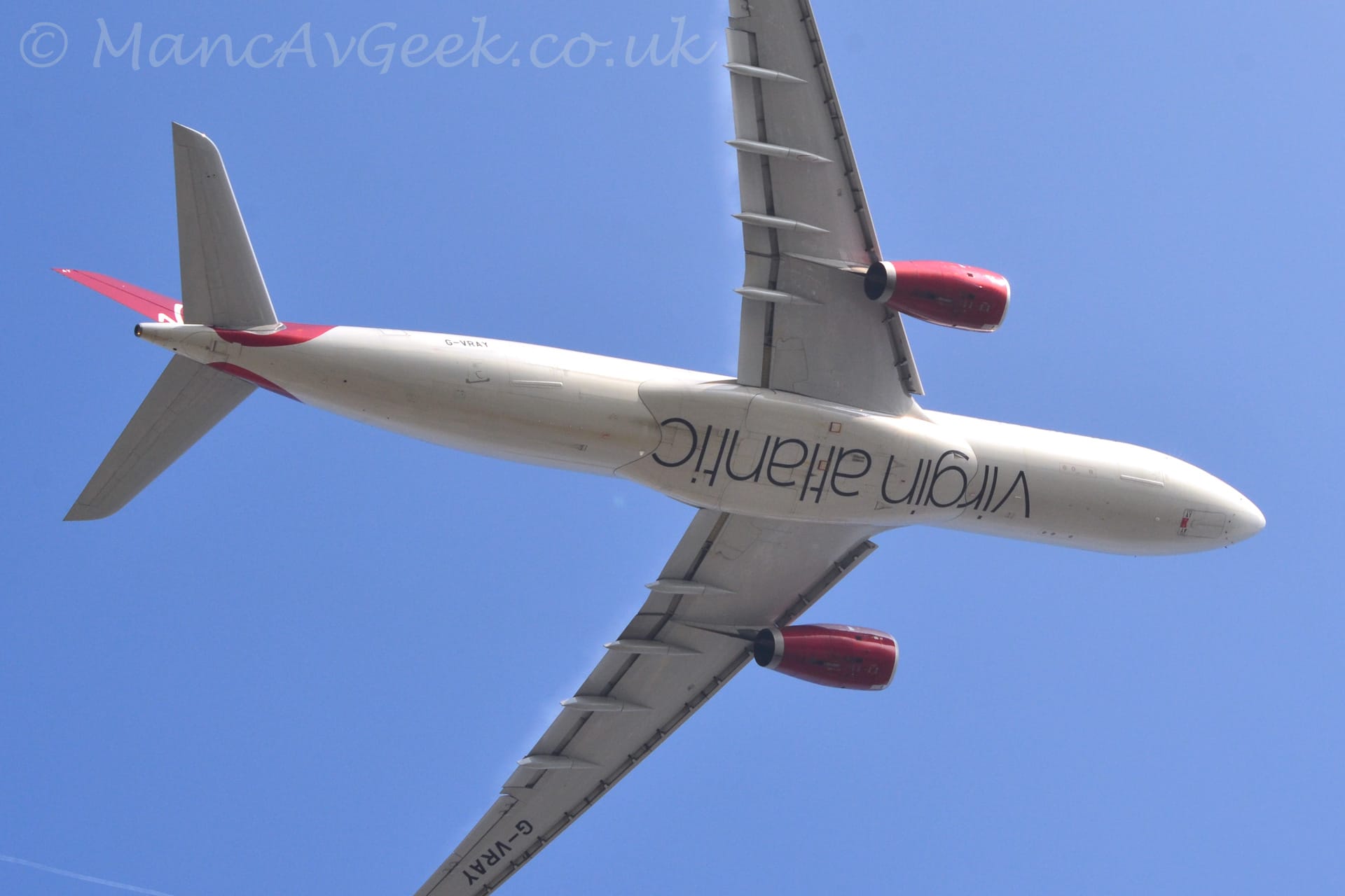 Low view of a twin engined jet airliner flying from left to right almost directly overhead at a very low altitude. The undercarriage has already been tucked away, and flaps are deployed from the rear of the wings, with the nose raised sharply, suggesting it has just taken off. The plane is almost entirely white, with black inverted "Virgin Atlantic" titles on the wingbox under the middle of the fuselage. The tail and the engine pods under the wings are a bright red. The black registration "G-VRAY" is visible on the lower rear fuselage, as well as under the outer section of the left wing. Blue sky fills the rest of the frame.