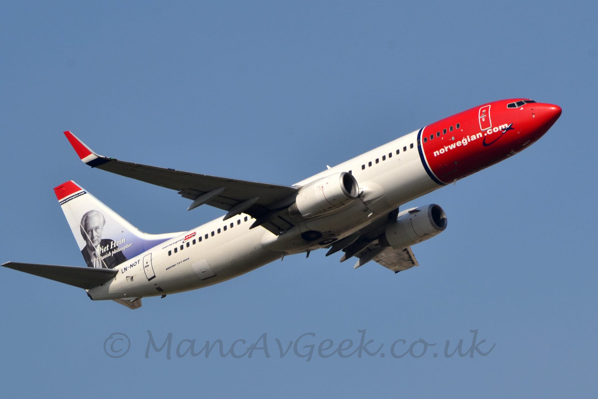 Low side view of a twin engined jet airliner flying from left to right at a very low altitude. The undercarriage has been tucked away, flaps have been extended from behind the wings, and the nose is raised sharply, suggesting it has just taken off. The plane is mostly white, with the nose and forward fuselage being red, with a thin blue band separating that from the white. The white web address "norwegian.com" is on the lower section of the red forward fuselage. The tail has a thick red and thin blue stripe at the top, with a black and white image of a man filling most of the rest, overlaid with the white text "Piet Hein". Blue sky fills the rest of the frame.