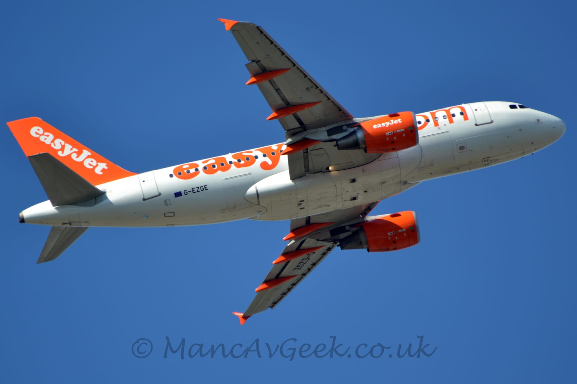 View from below of a twin engined jet airliner flying from left to right almost overhead, with flaps extended from the rear of the wings, undercarriage stowed away, and nose raised sharply, suggesting it has just taken off. The plane is mostly white, with orange billboard-style "EasyJet.com" titles on the fuselage, and white diagonal "EasyJet" titles on the orange tail, as well as the orange engine pods mounted under the wings. There are orange covers on the tracks that move the flaps under the rear of the wings, and the vertical plates on the wing-tips are also orange. The black registration "G-EZGE" is on the lower rear fuselage, next to a small EU flag, as well as under the left wing. Vivid blue sky fills the rest of the frame.