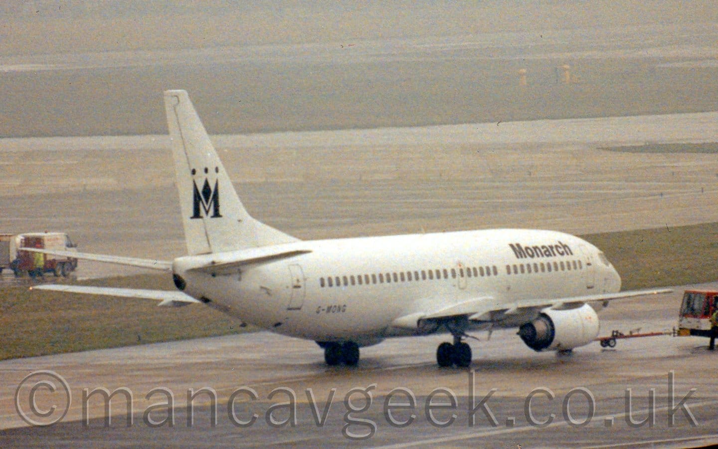 Side view of a twin engined jet airliner, facing to the right and slightly away from the camera but being pushed back to the left by a low-slung red and white tug attached to the planes nose-wheel by a long red pole. The plane is almost entirely white, with black "Monarch" titles on the upper forward fuselage, the black registration "G-MONG" on the lower rear fuselage, and black crown that resembles an upper-case letter "M" on the tail. Grey and black apron fills the foreground, with a strip of green grass behind the plane separating it from a pair of taxiways, more grass, and a runway in the background, slowly vanishing in to haze.