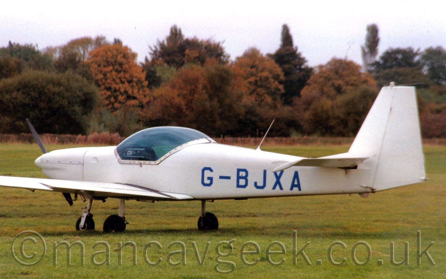 Side view of a single-engined light aircraft parked on grass facing to the left. The plane is mostly white, with blue registration "G-BJXA" on the rear fuselage. A large, blue-tinted bubble canopy covers the cockpit, over the wing root. Grass fills the foreground and most of the background, with green and brown trees in the distance, under grey sky.