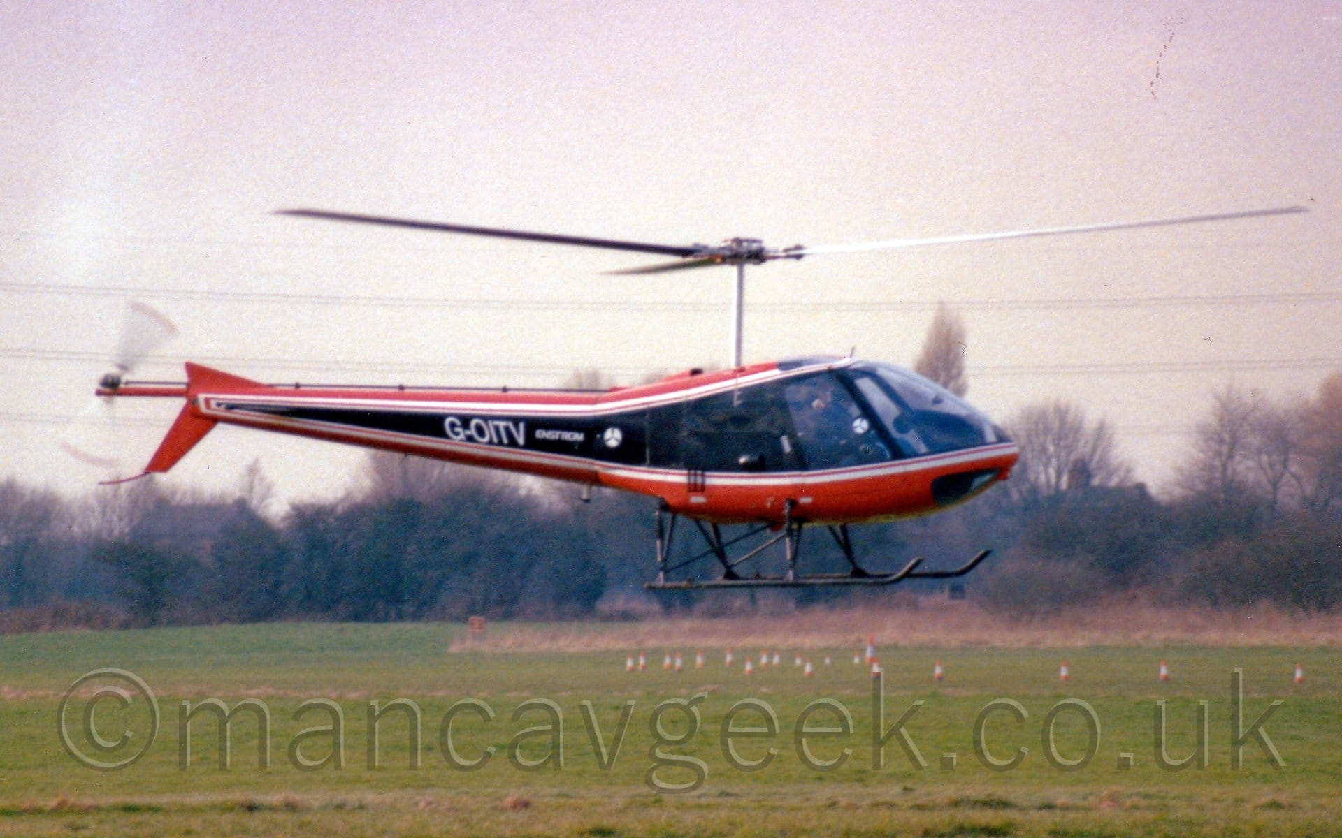 Side view of a light helicopter hovering 3 or 4 metres above a grass airfield. The 'copter has a wide black stripe running along the body, narrowing slightly as it moves to the rear of the tail boom, outlined in white, with the upper and lower surfaces being an orange-brown. The white registration "G-OITV" is on the tailboom, next to a small "Enstrom" logo. Green grass fills the bottom of the frame, with trees in the distance, while grey sky fills the top.