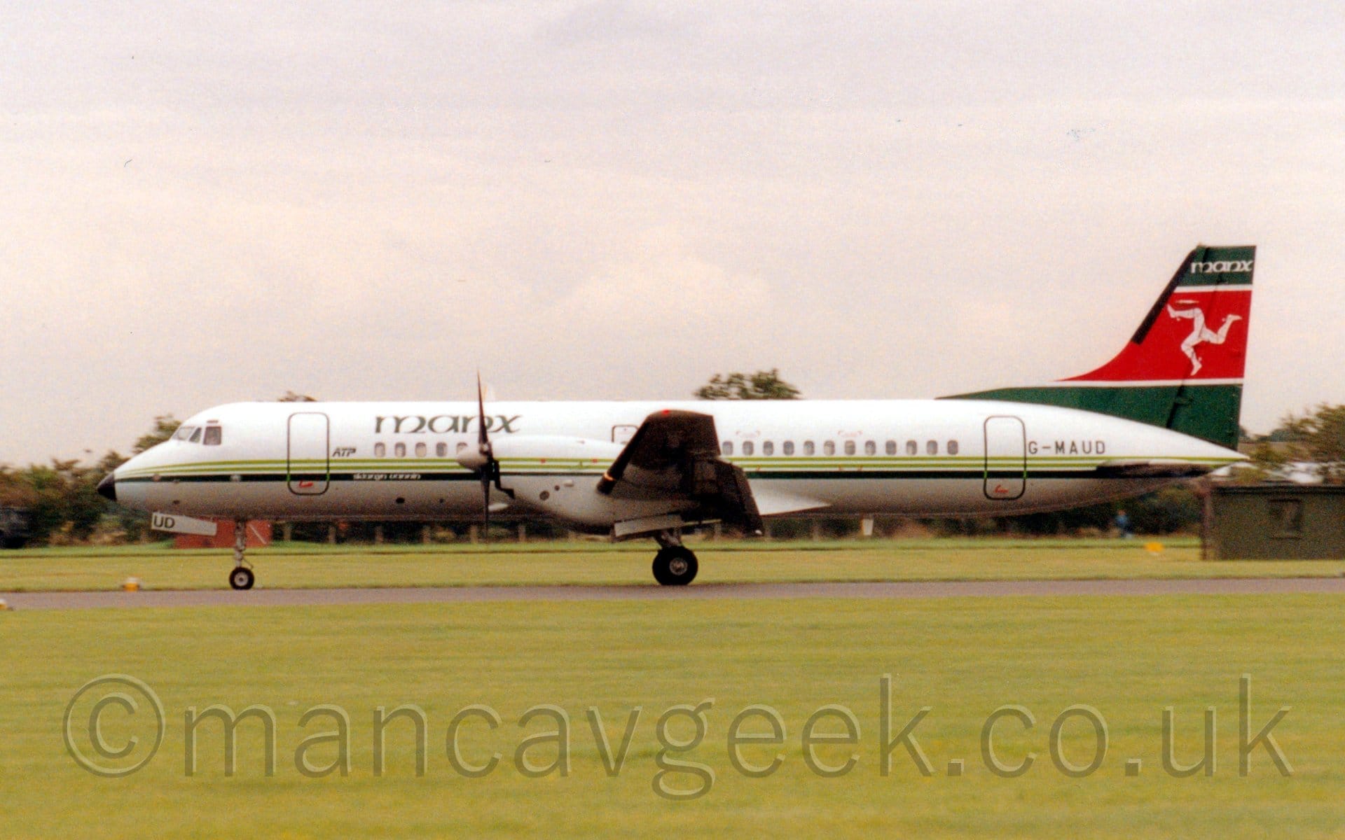 Side view of a twin propellor-engined airliner taxiing from right to left along a black runway. The plane is mostly white, with 2 thin light green stripes and a slightly thicker dark green stripe running along the body, with dark green "Manx" titles on the upper forward fuselage, green "ATP" titles just aft of the forward door, and the green registration "G-MAUD" just aft of the rear door. The tail is the same dark green, with a thick red band outlined in white, with a white triskeles made up of 3 armoured legs with spurs on the heels, joined at the hip, and seeming to run in a circle to the right. The top of the tail has white "Manx" titles at the top. Lush green grass fills the foreground, with more grass visible under the plane, leading up to trees in the distance, under grey sky.