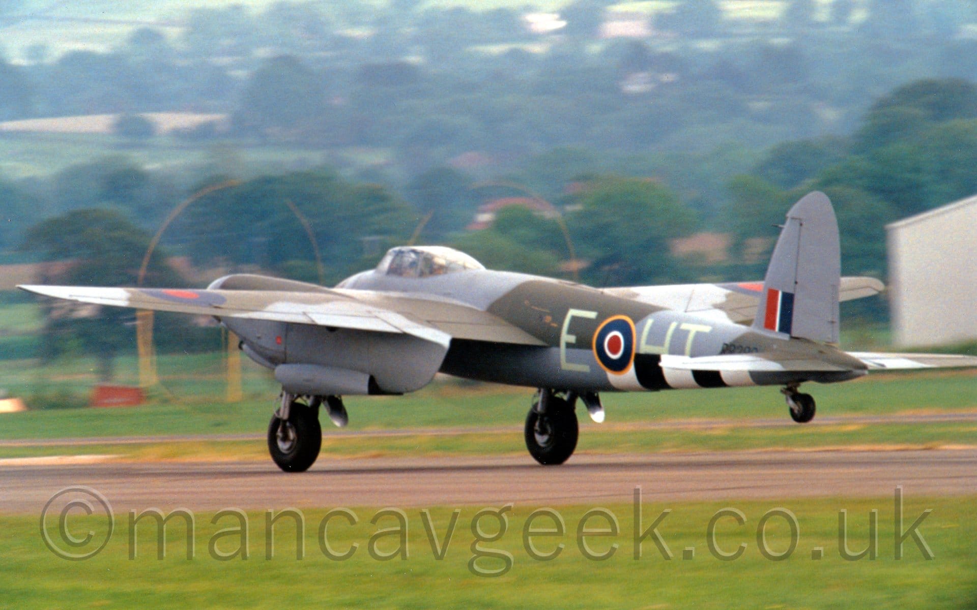 Side view of a twin propellor-engined, World War 2-era fighter-bomber accelerating from right to left along a grey tarmac runway, with it's tail wheel raised off the ground, suggesting it is taking off. The plane has a grey and olive green camouflage scheme on the upper body and upper wing surfaces, with grey belly and undersides of the wings. There are 3 white and 2 black bands under the lower rear fuselage. The pale mint green letters "E-HT" are either side of a blue, white, and red roundel, outlined in yellow, on the rear fuselage, with the black serial "RR299" just after, under a red, white, and blue fin-flash on the tail. Green grass fills the foreground, with trees on small hills in the background.