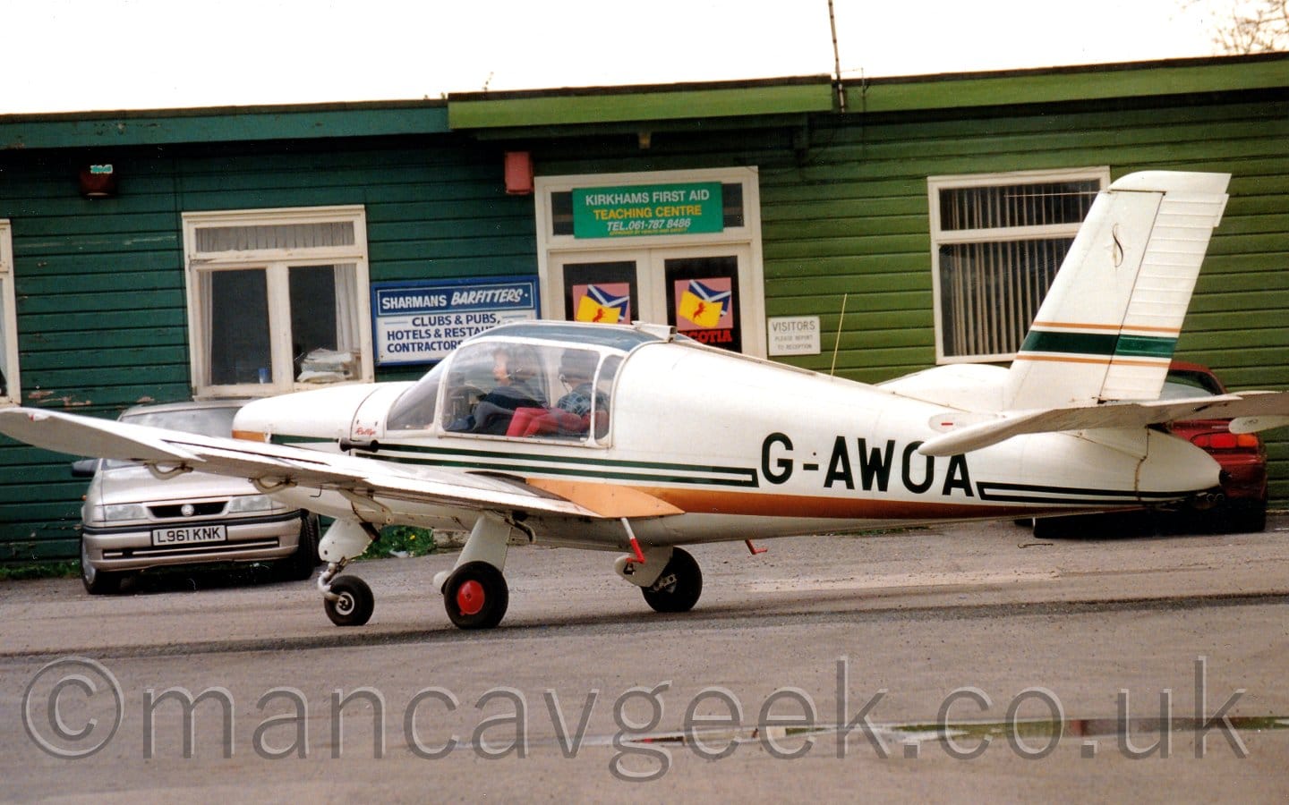 Side view of a single engined light aircraft taxiing from right to left on a tarmac apron. The plane is largely white, with a dark blue and gold stripe running along the body, with the black registration "G-AWOA" partially overlaid on the rear fuselage. A large glazed canopy covers the cockpit, and the 2 occupants can be seen. A green wooden building with a car parked in front stretches across the background