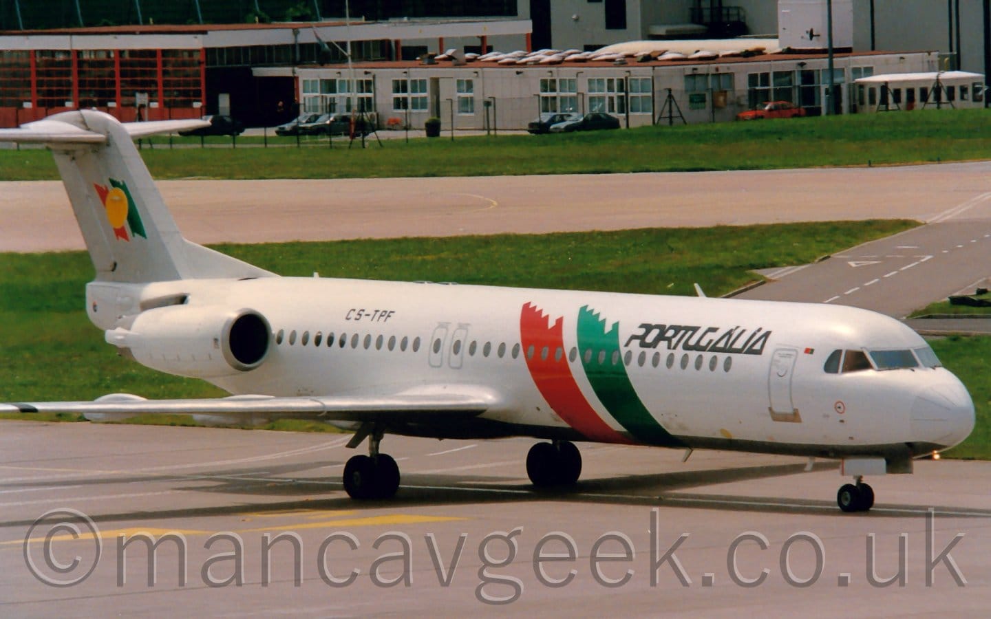 Side view of a twin engined jet airliner taxiing from left to right. The plane is mostly white with a grey belly and black "Portugalia" titles on the upper forward fuselage. The black registration "CS-TPF" is on the upper rear fuselage, in front of the engines which are mounted on the sides of the rear fuselage. A pair of diagonal stripes, one red, one green, are on the side of the side of the forward fuselage, in front of the wings. Smaller versions of these stripes appear on the grey tail, overlaid with a yellow circle.  Grey taxiway fills the foreground, with a wide strip of grass in the background separating it from another taxiway, with more grass and a low cream-coloured building beyond that.