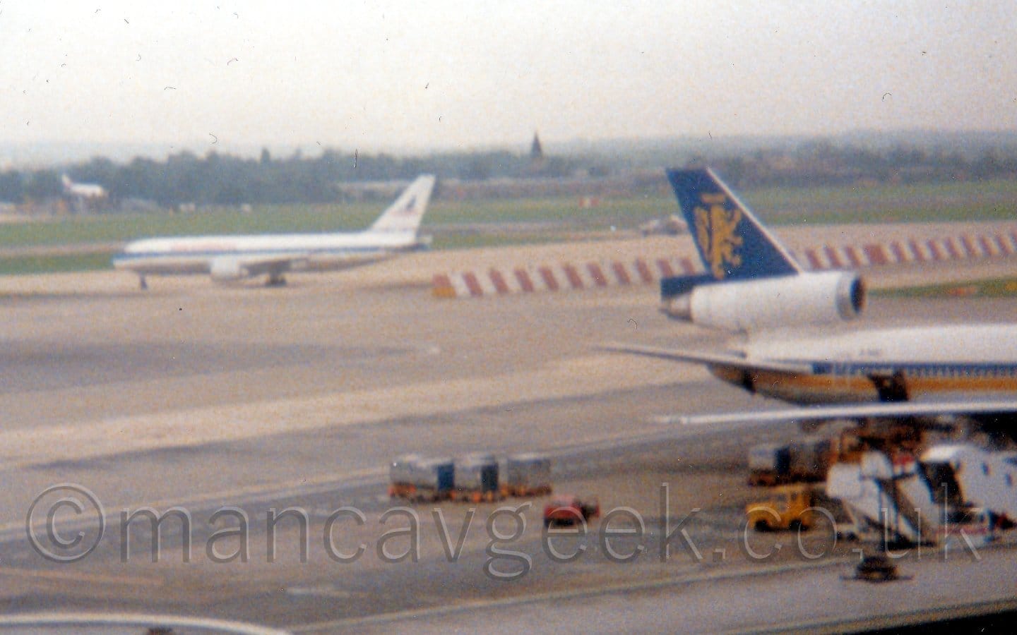 Rather grainy shot of a white, twin engined jet airliner taxiing from right to left in the distance, with the rear fuselage and tail of a 3 engined jet airliner on the right of the frame. The twin engined plane has a blue stripe running along the body, the other has a yellow and blue body stripe, with a blue tail containing a yellow lion standing on hits hind legs. A large expanse of grey apron fills most of the frame, with grass in the background leading up to trees in the distance, under hazy grey sky.