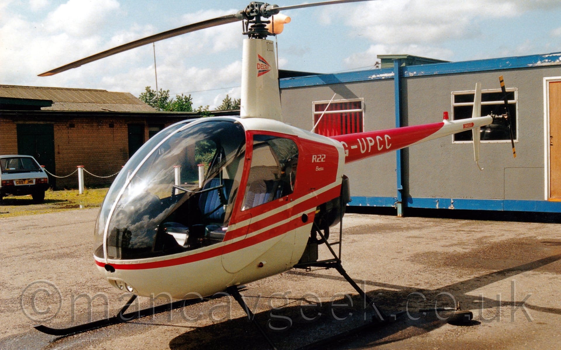Side view of a light helicopter parked on grey concrete facing to the left. The chopper is mostly white, with red trim around the cockpit and on the tail boom. The tail boom has the white registration "G-UPCC" on the red section. A grey prefabricated building with a blue frame is in the background on the right, while a brown brick building with a grey car parked in front is on the left.