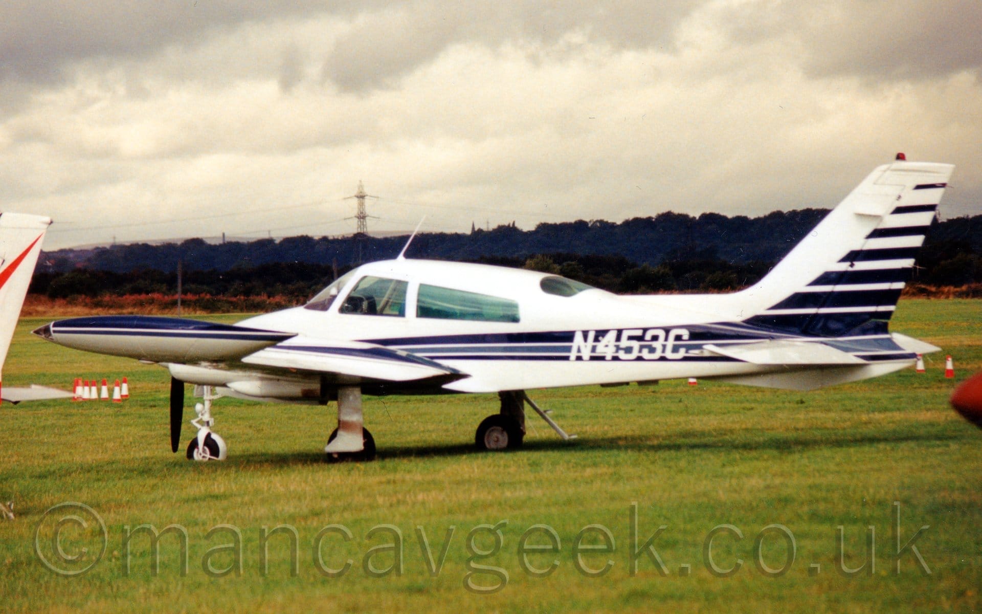 Side view of a twin engined light aircraft parked on grass facing to the left. The plane is mostly white, with 3 blue stripes running along the body, getting wider as they move rearwards, where they are joined by more stripes at the rear of the tail. The white registration "N453C" is overlaid on the stripes on the rear fuselage. There are more blue stripes on the top of the engine cowlings on top of the wings, as well as on the wing-tip fuel tanks. Green grass fills the foreground, with the red nosecone of another plane on the right of the frame, and the white and red tail on the left. Trees line the airfield perimeter in the distance, with rolling hills beyond that, and an electricity pylon looming over everything, under moody, cloudy grey sky.