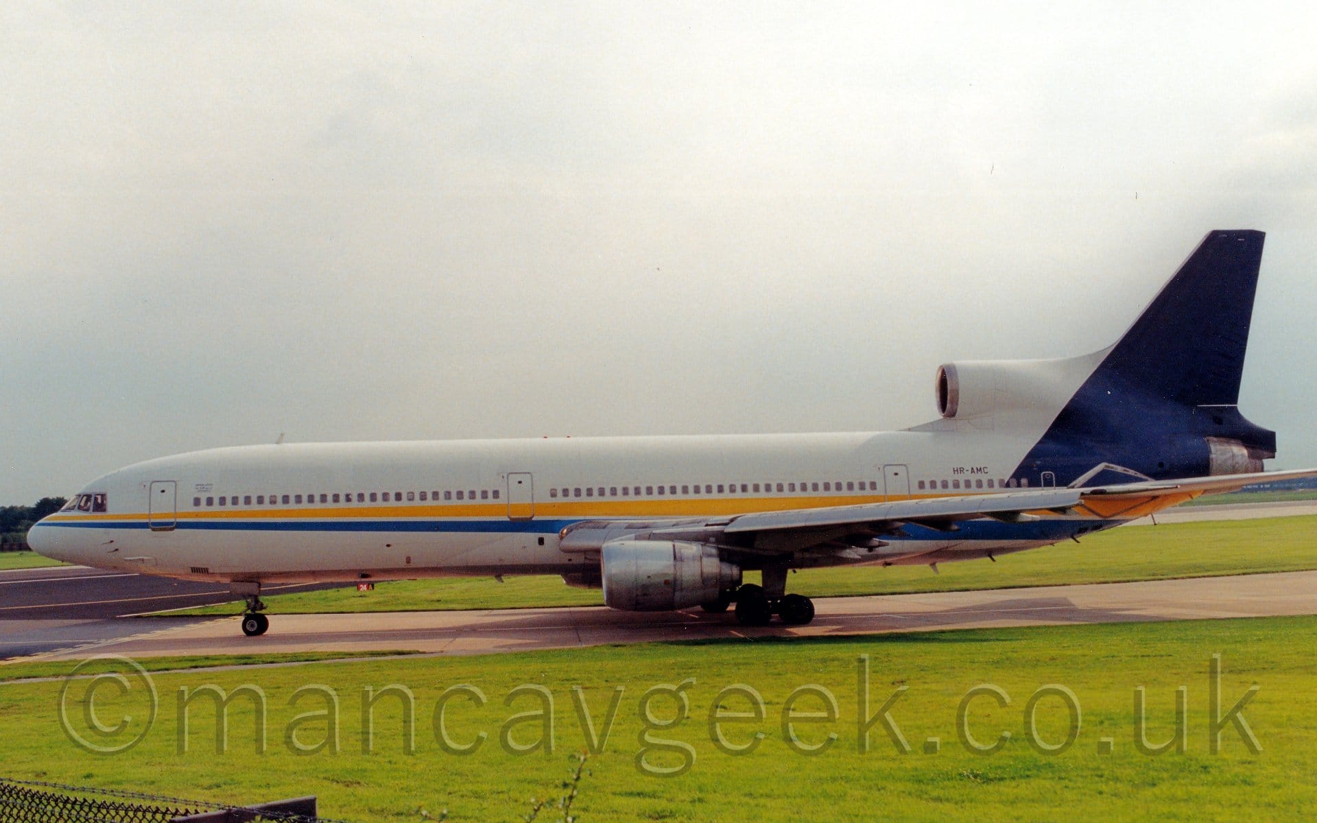 Side view of a 3 engined jet airliner taxiing from right to left along a grey taxiway, about to turn on to a black runway. The plane is mostly white, with a yellow and dark blue stripe running along the body from the nose, getting slightly wider as it moves rearwards, as well as a dark blue tail. There are no other markings, apart from some very small, indecipherable text by the forward cabin door, and the black registration "HR-AMC" on the rear fuselage, under the centre engine intake. Lush green grass fills the foreground, with more grass visible under the plane in the background. Flat grey sky with a bright spot right at the top fill the rest of the frame.