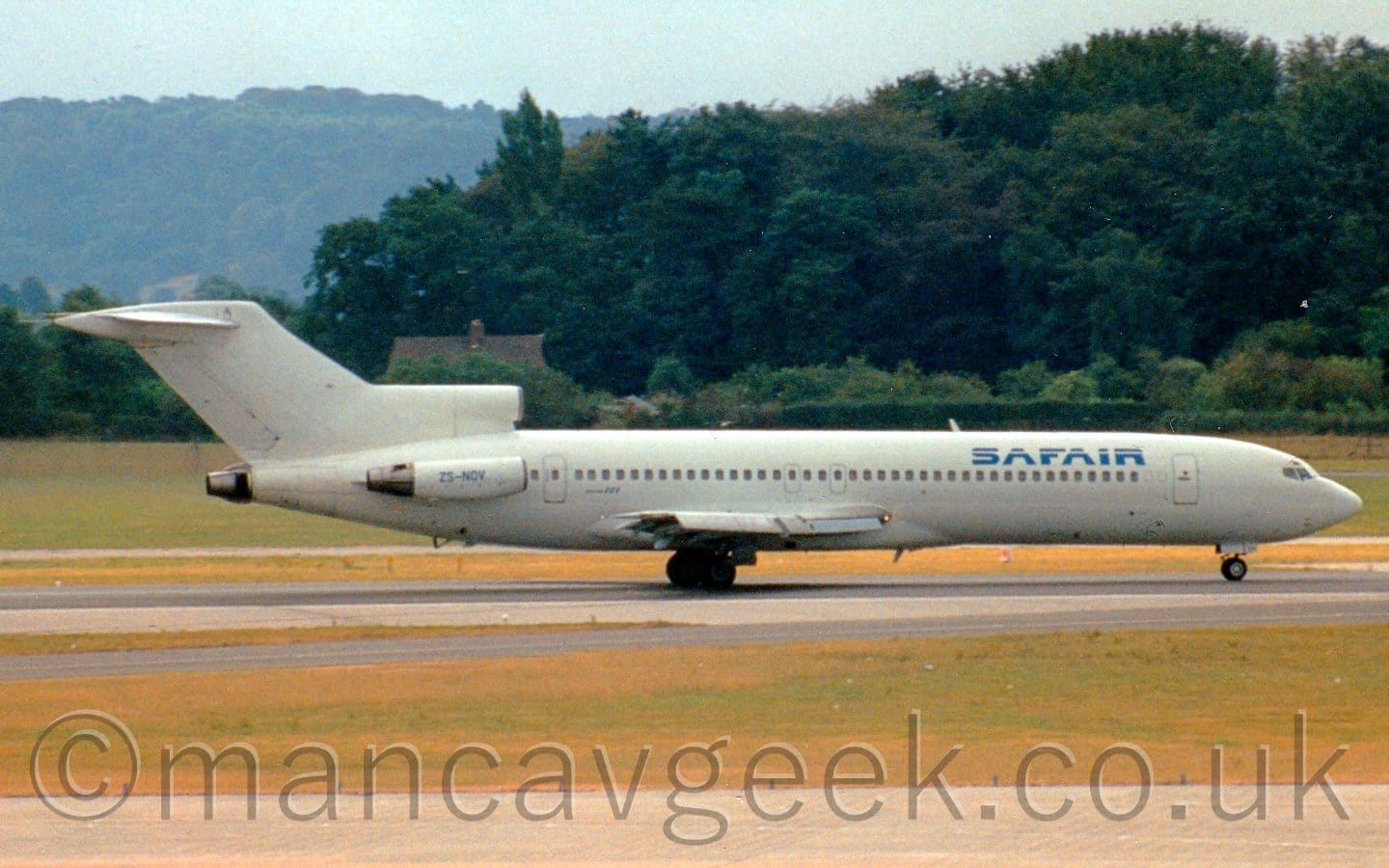Side view of a white, 3 engined jet airliner moving from left to right, decelerating hard after landing. The plane is almost entirely white, with only blue "SafAir" titles on the upper forward fuselage, and the registration "ZS-NOV" on the sides of the engine pods mounted on the sides of the rear fuselage adding a splash of colour. Flaps extend from the front and rear of the wing, suggesting it is slowing down in a hurry after landing. Large grassed areas mixed with grey taxiways fill the foreground, with a lonely house nestled in trees in the background. Trees on a distant hillside and grey sky fills the rest of the frame.
