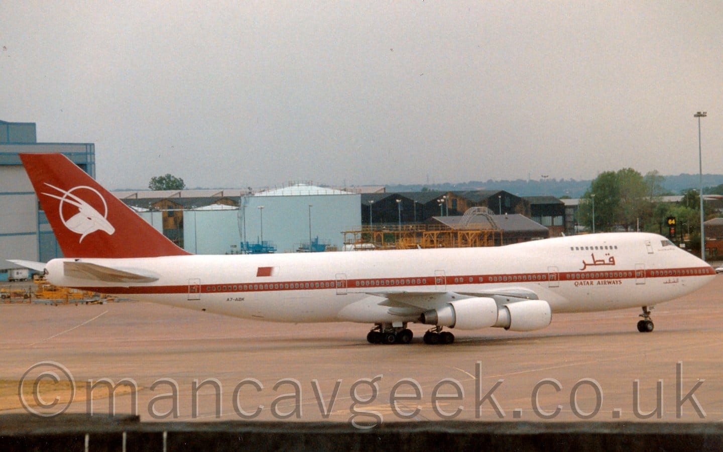Side view of a very large, 4 engined jet airliner taxiing from left to right. The plane is mostly white, with a maroon stripe running along the body, covering the lower deck windows, and maroon "Qatar Airways" titles on the forward fuselage, in an Arabic script between the upper and lower deck windows, and in English below the lower deck windows. The tail is also maroon, with the white head of a horned, goat-like animal inside the outline of a white circle. Grey concrete apron fills the foreground, with a large grey hangar in the background on the left, several pale blue fuel tanks towards the middle, and a selection of other airport buildings on the right. Trees in the distance slowly vanish into a bright but hazy sky.