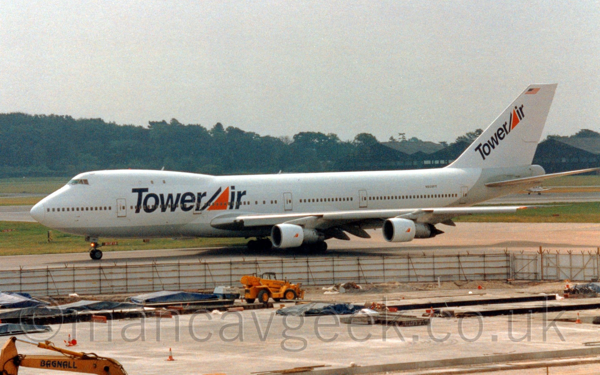 Side view of a very large, 4 engined jet airliner taxiing from right to left. The plane is mostly white, with a grey belly, and black "TowerAir" titles on the side of the forward fuselage, the letter "A" represented by a black diagonal line followed by an orange right-angled triangle. This is repeated diagonally on the tail, with the black diagonal line and orange triangle used as a logo on the sides of the white engine pods slung under the wings. There is also a small US flag at the top of the tail, and the black registration "N609FF" on the upper rear fuselage. A fenced-off building site fills the foreground, with trees stretching across the background from the left, and a series of black hangars on the right of the frame, with flat grey sky over everything.